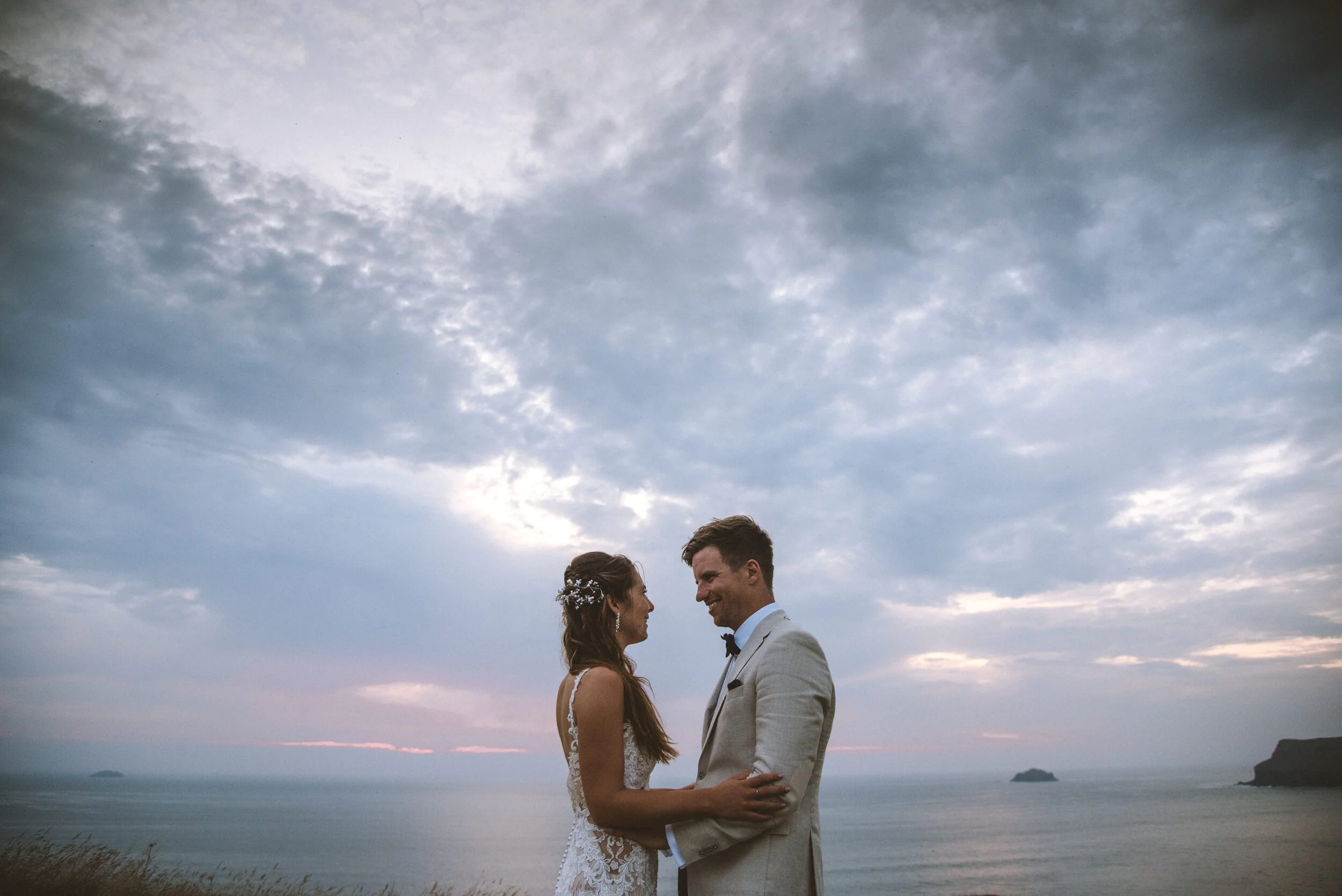 Daymer bay wedding photography in Cornwall captured by Mark Shaw Photography - A bride and groom in wedding attire standing outdoors near the ocean, facing each other and smiling under a cloudy sky.