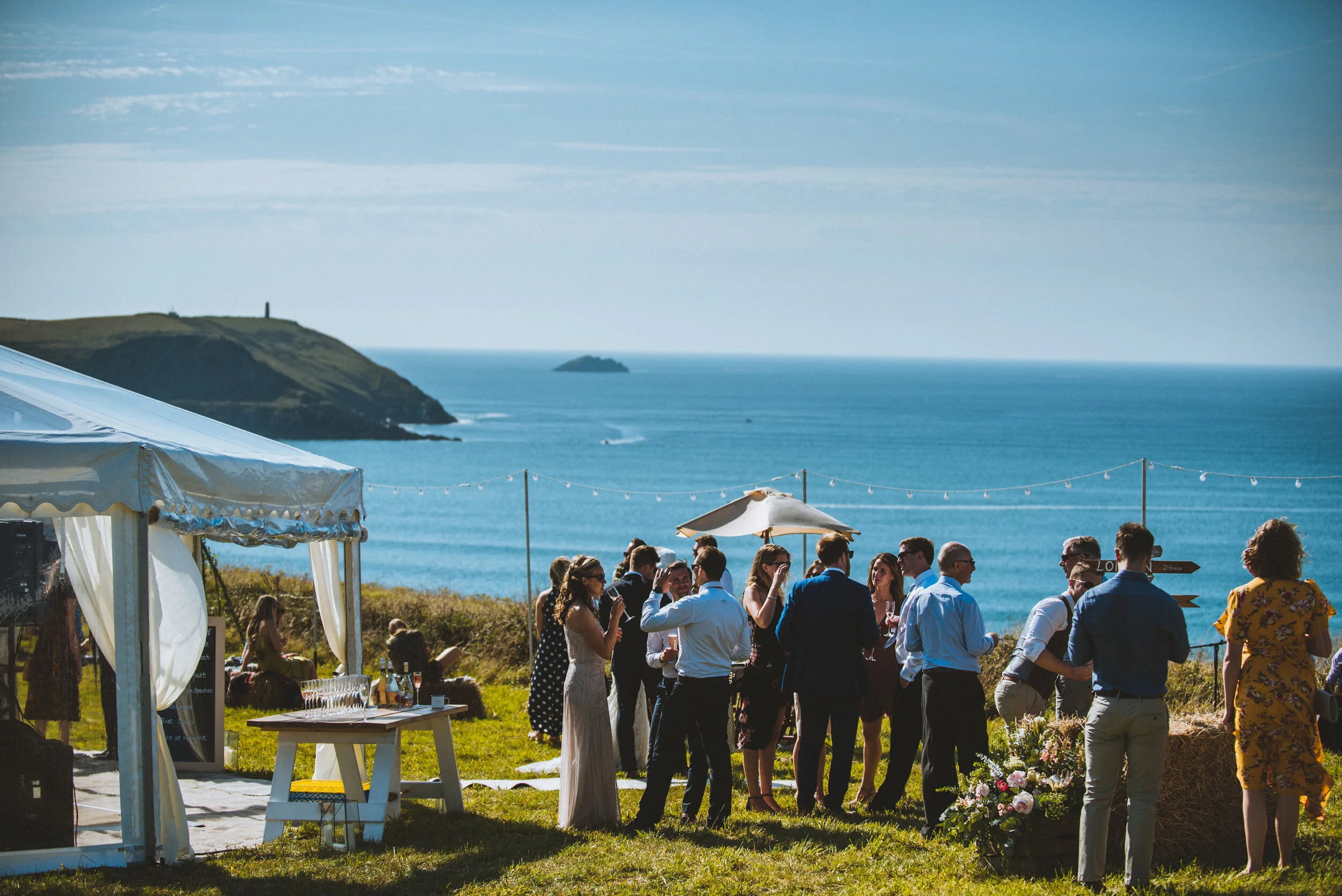 Isles of scilly photography. People gathering outdoors at a seaside event, with a tent, string lights, and an ocean view in the background.