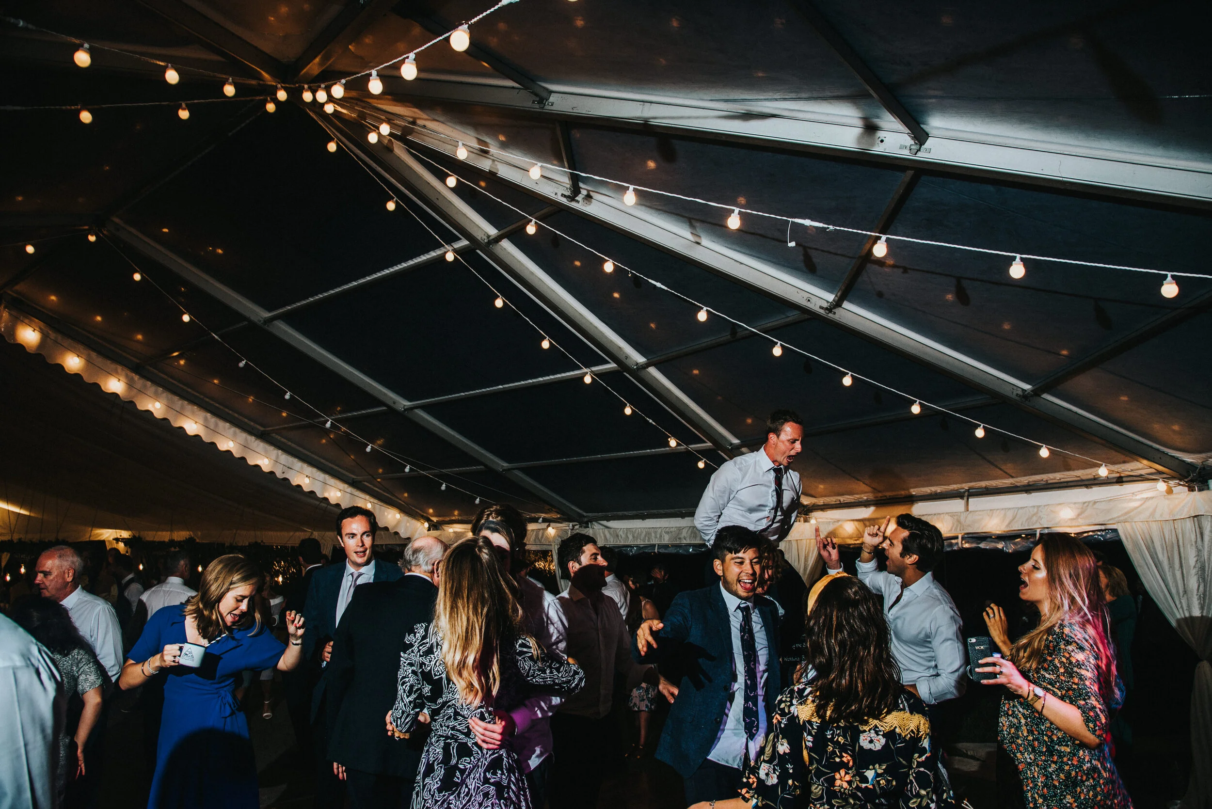 Daymer bay wedding photography captured by Cornwall wedding photographer Mark Shaw Photography -People dancing and celebrating at a party under a decorated tent with string lights.
