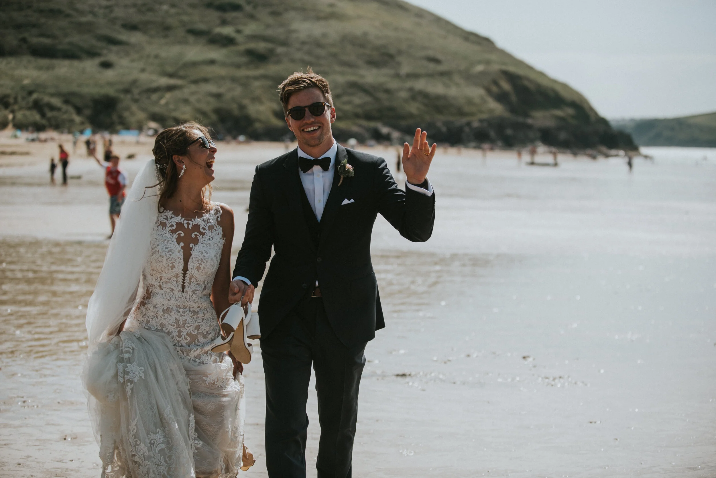 A bride and groom walking on a beach, smiling and laughing, with the bride holding her shoes in her hand and the groom waving. The bride is wearing a lace wedding dress and veil, and the groom is in a black tuxedo with sunglasses. There are people an