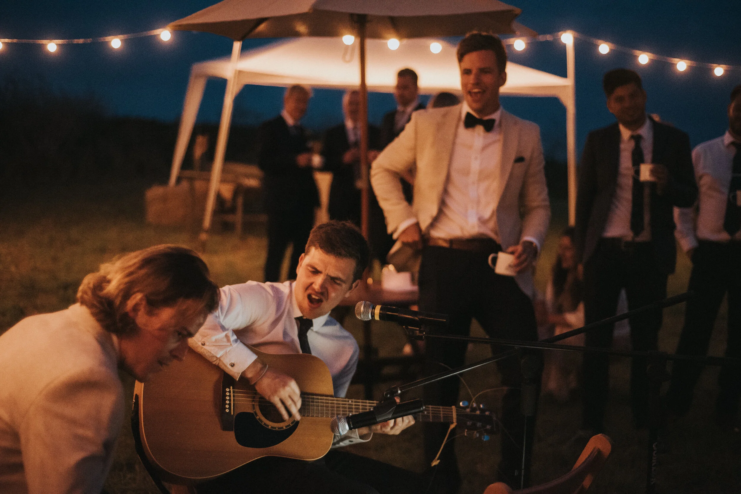 Daymer bay wedding photography captured by Cornwall wedding photographer Mark Shaw Photography - People in formal attire enjoying live acoustic music during an outdoor evening event, with string lights illuminating the scene.