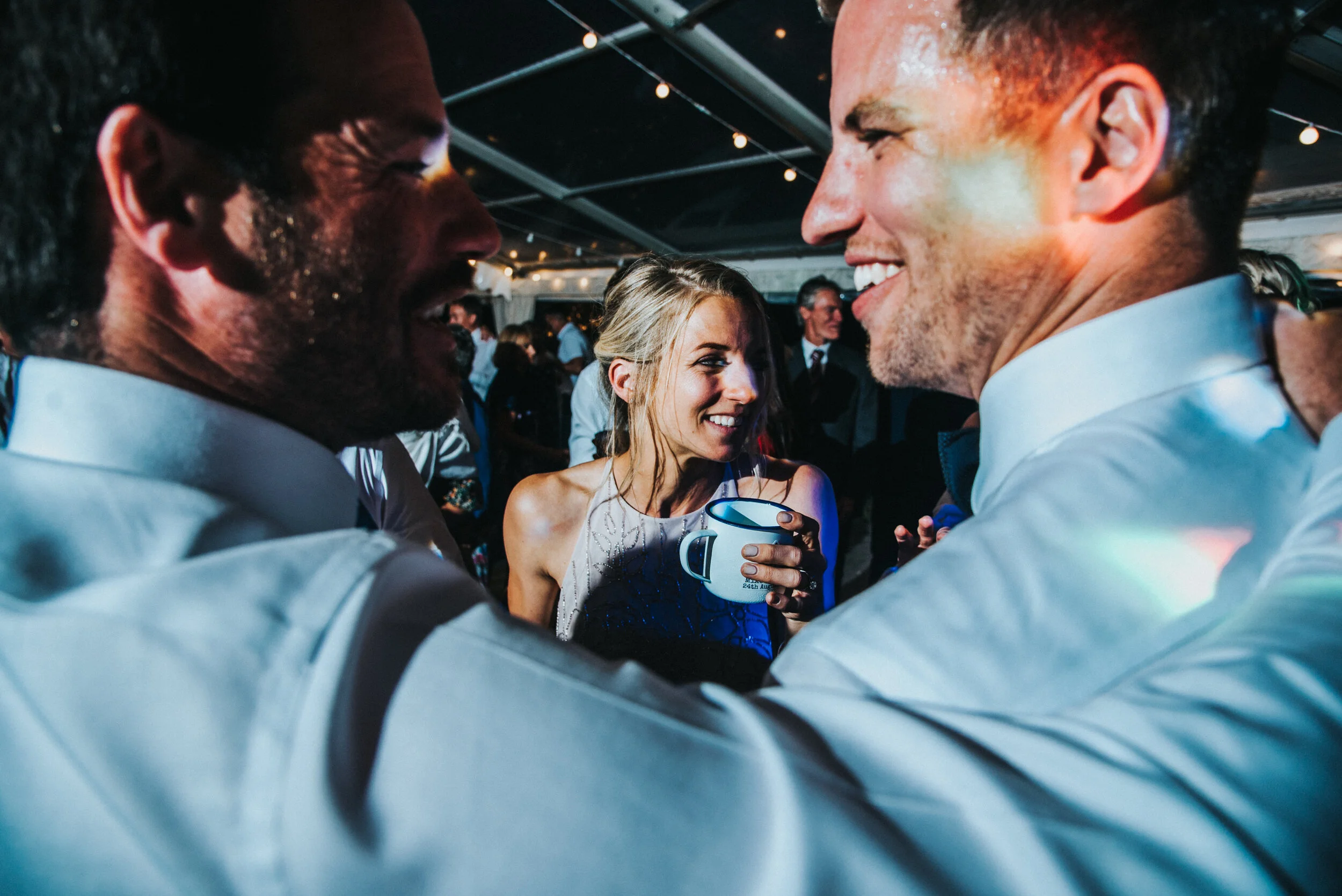 Two men in tuxedos smiling and facing each other, with a woman in a sleeveless dress holding a mug and smiling in the background at a lively indoor event.