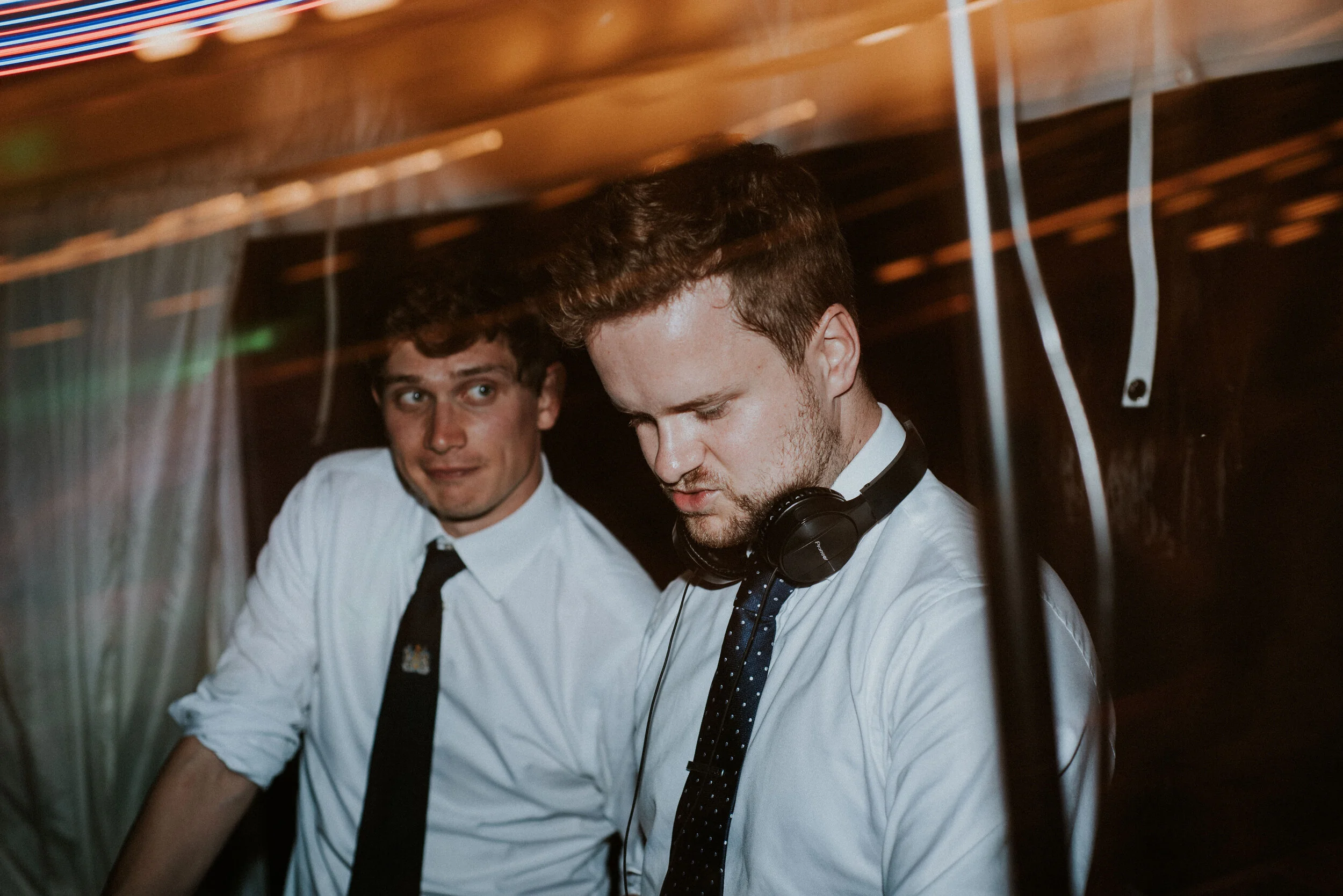 Daymer bay wedding photography captured by Cornwall wedding photographeTwo young men wearing white shirts and black ties, one with headphones around his neck, standing in a dimly lit environment with colorful lights, possibly a DJ booth or nightclub.