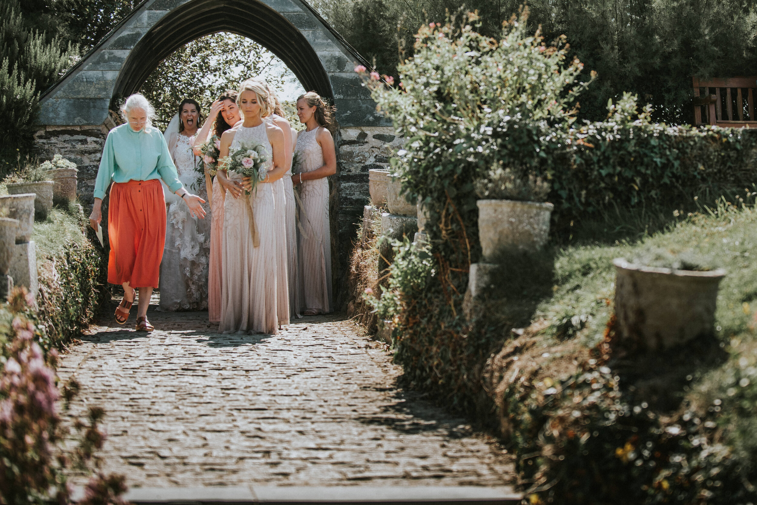 Daymer bay wedding photography in Cornwall captured by Mark Shaw Photography - Group of women walking under an archway on a stone pathway, with women in bridesmaid dresses and one holding a bouquet, surrounded by greenery and flower pots.