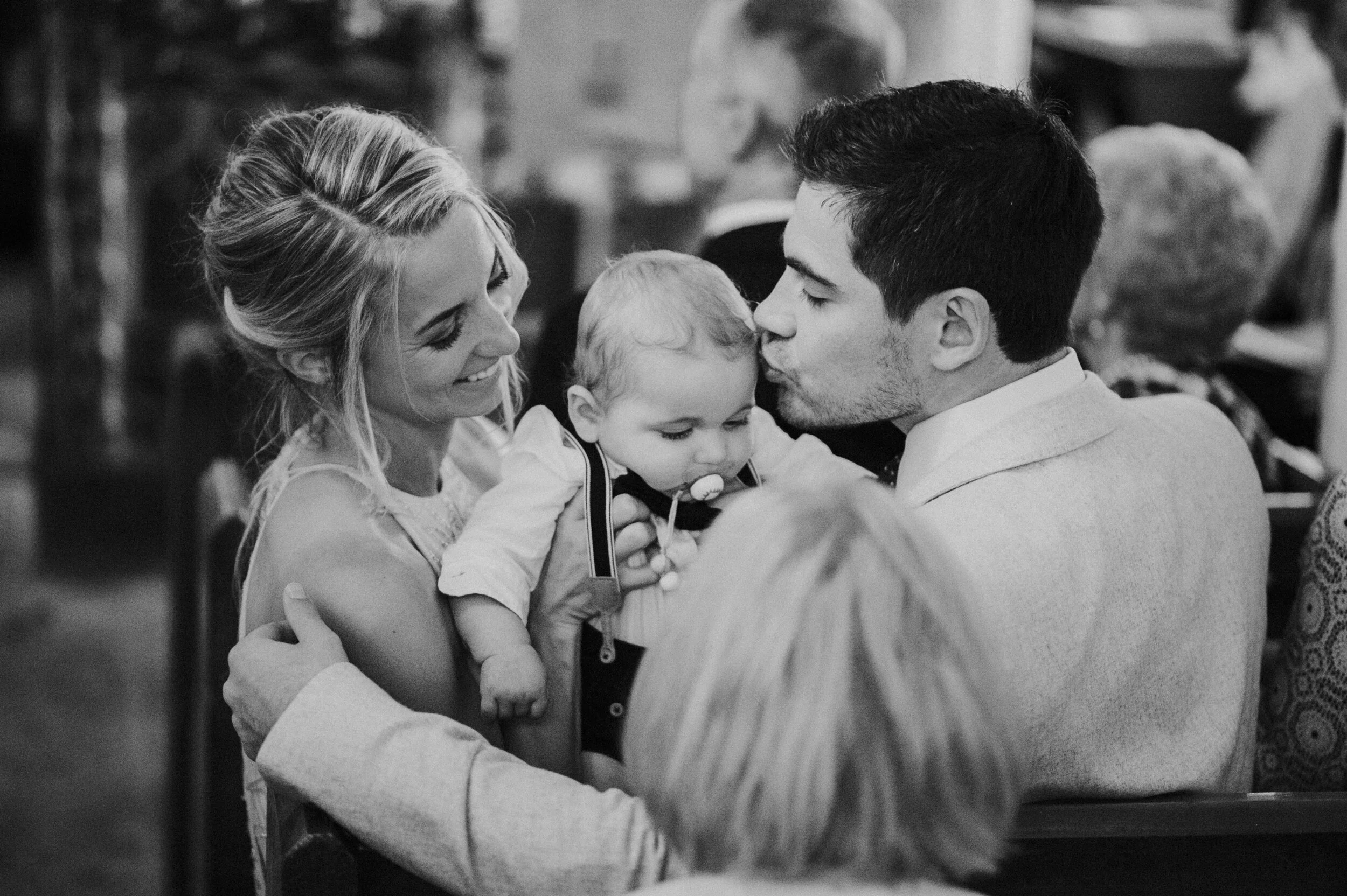 A black and white photo of a family at a gathering. A woman and a man hold a young child between them. The man kisses the child on the forehead, and the woman smiles.