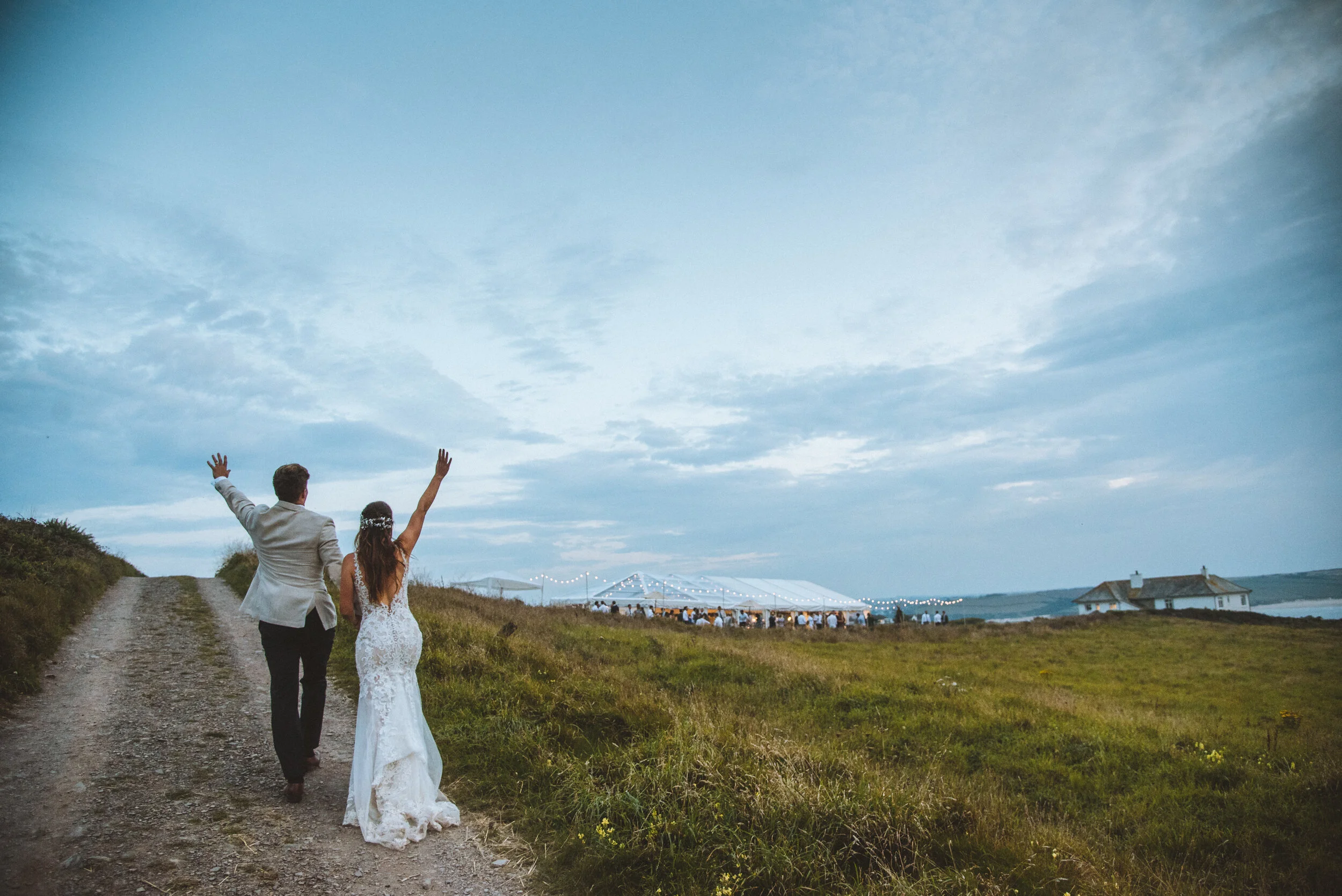 Daymer bay wedding photography captured by Cornwall wedding photographer Mark Shaw Photography - Bride and groom walking hand in hand on a dirt path towards a celebration tent in a rural landscape at dusk.