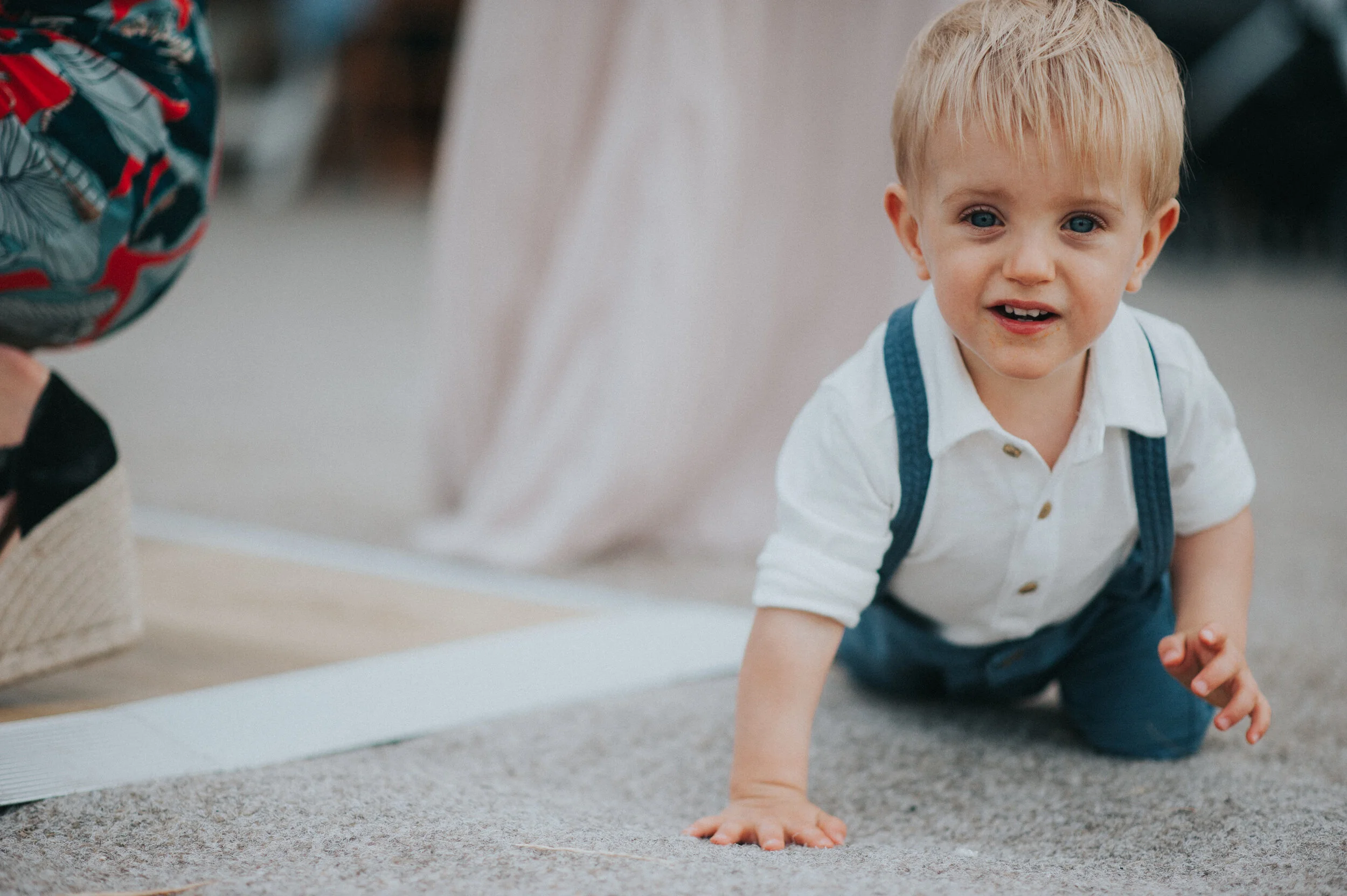 Daymer bay wedding photography in Cornwall captured by Mark Shaw Photography - A young boy crawling on the carpeted floor indoors, looking at the camera, wearing a white shirt and blue suspenders.