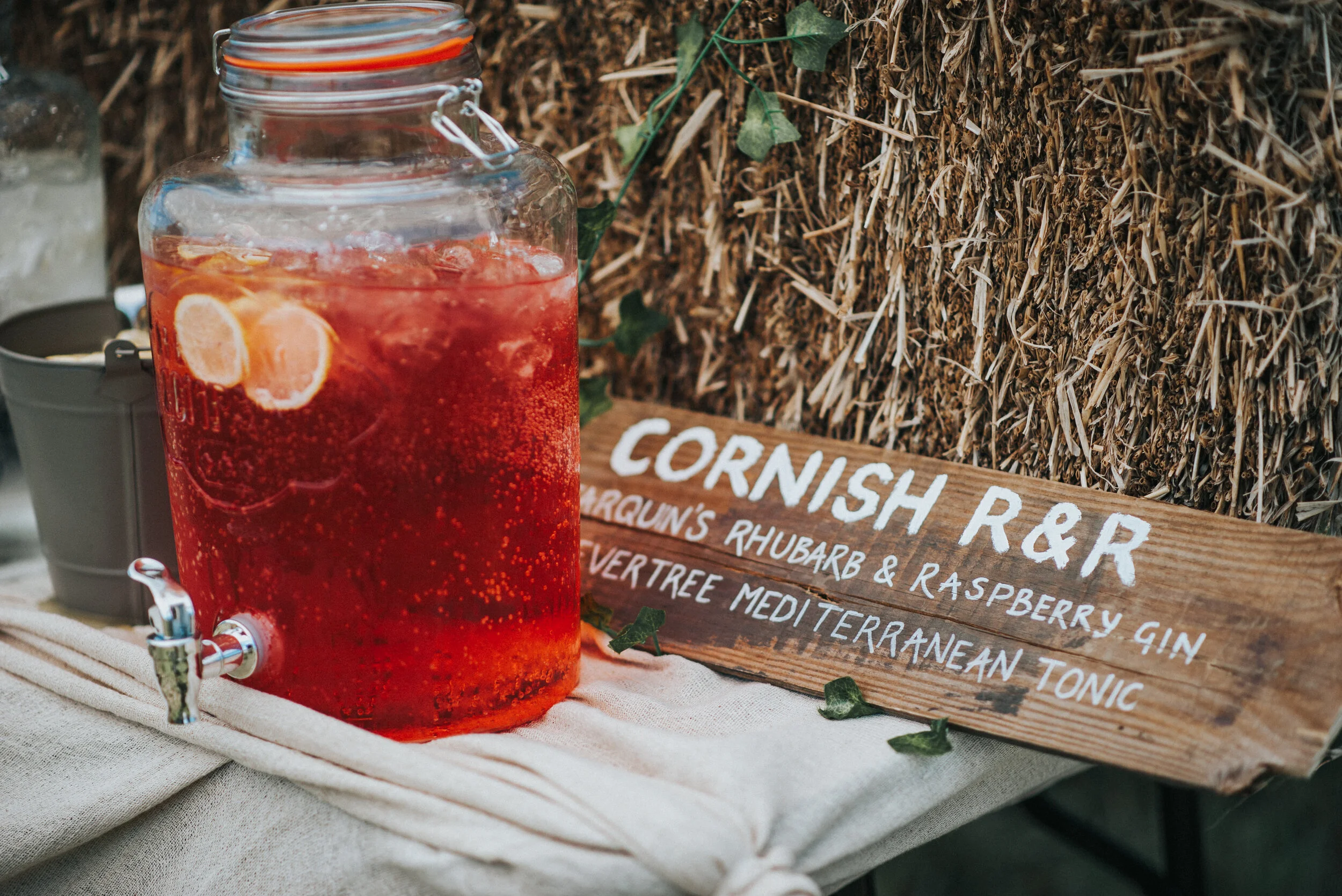 A large glass jar filled with a red, bubbly beverage garnished with lemon slices, sitting on a beige cloth table. Behind it, a wooden sign reads 'Cornish R & R,' and lists ingredients including rhubarb, raspberry gin, and Mediterranean tonic. The bac
