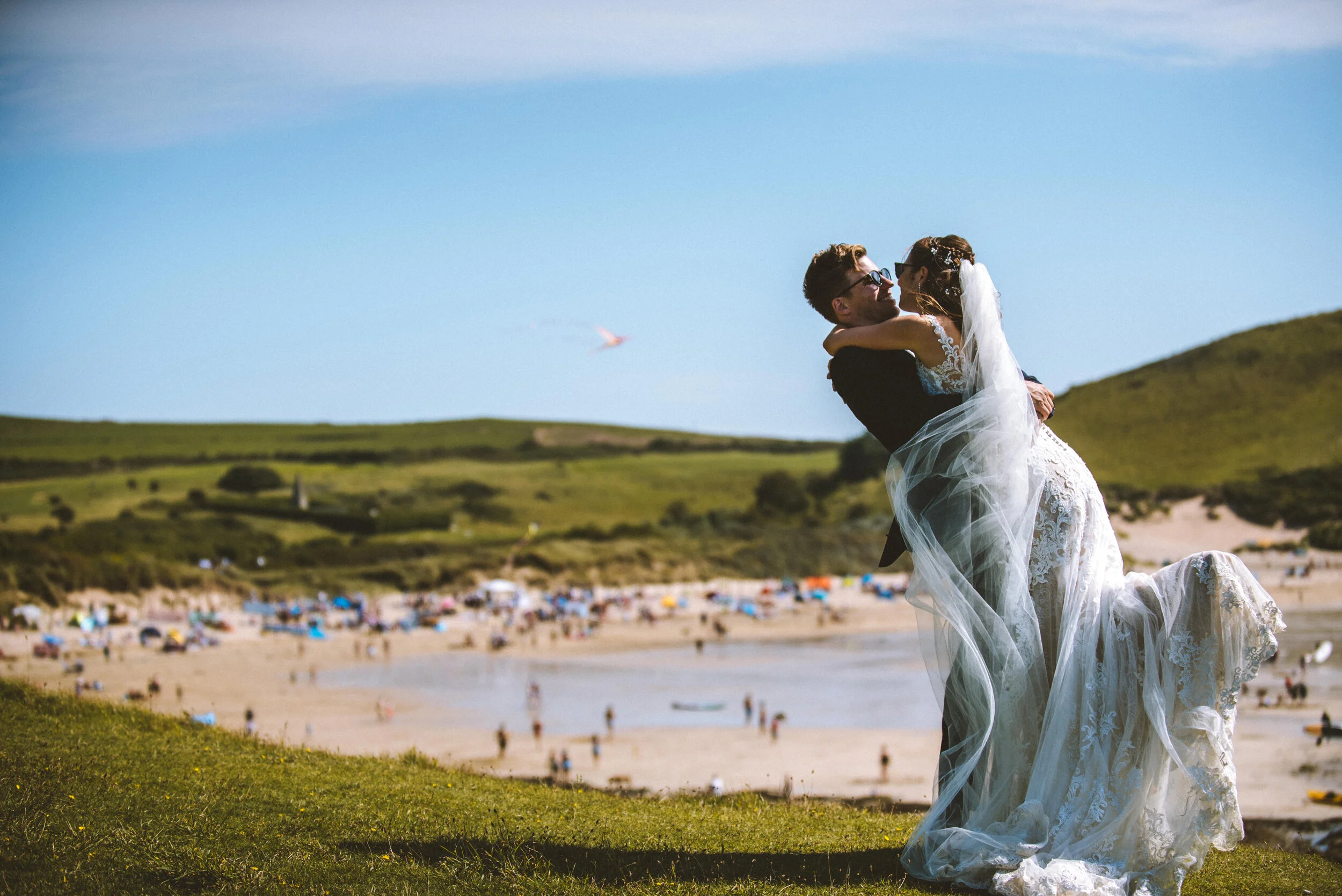 Isles of scilly photography. A newlywed couple dressed in wedding attire sharing a romantic moment on a grassy hill near a beach, with many beachgoers, umbrellas, and boats in the background.