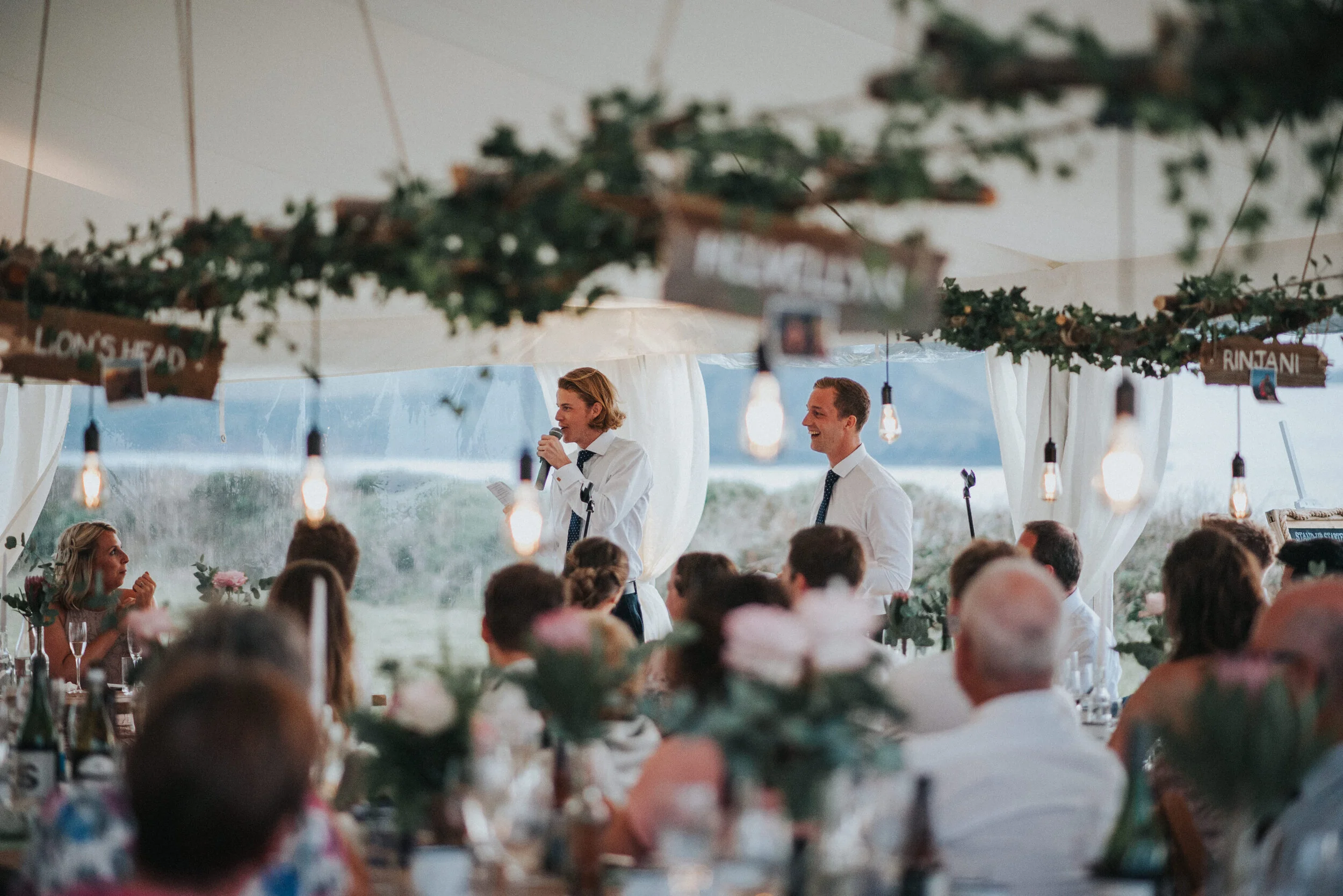 Daymer bay wedding photographA wedding reception under a tent with white drapery, decorated with greenery and hanging Edison bulbs, where two men in white shirts and black ties are giving speeches to seated guests, with a woman listening and smiling.