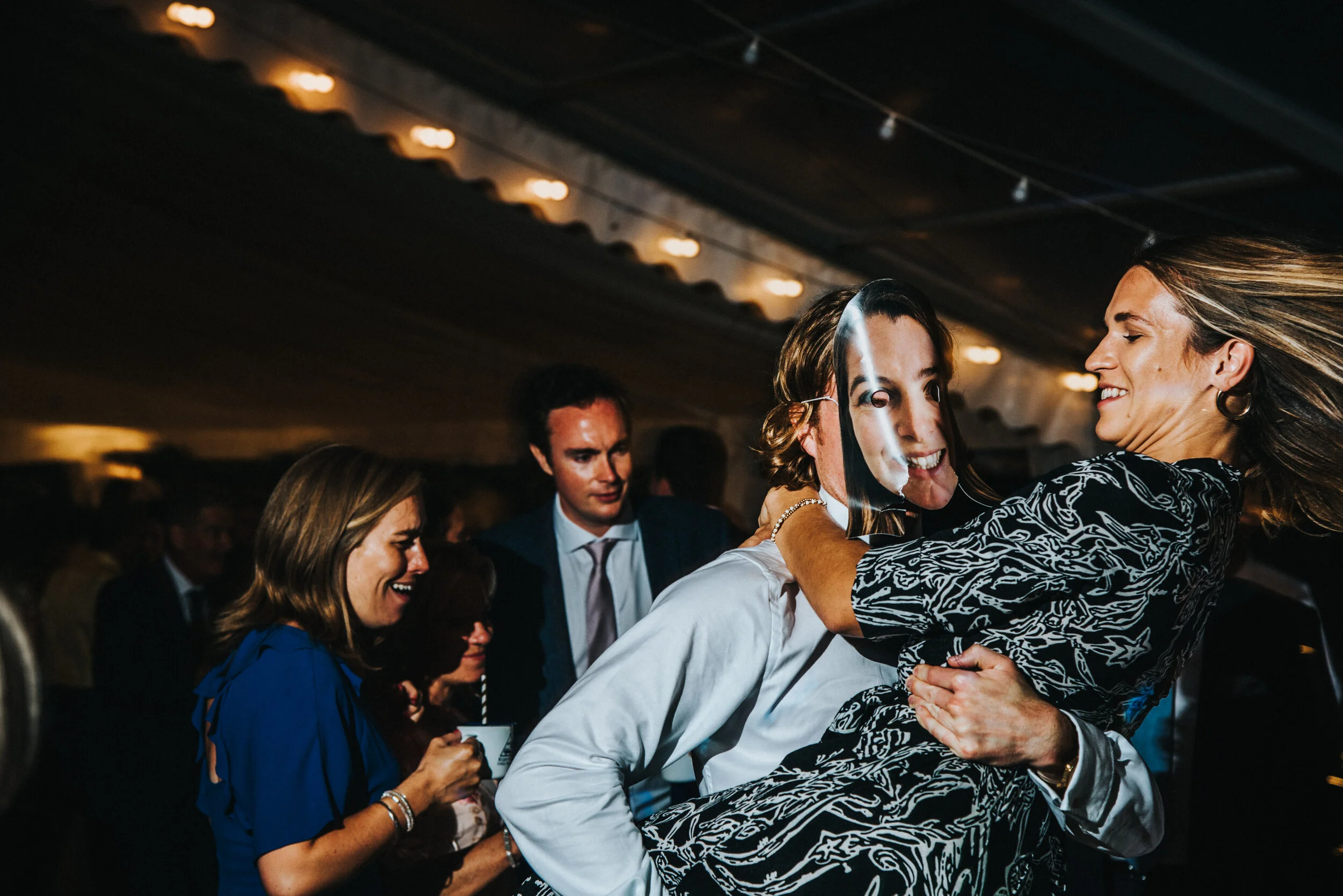 Daymer bay wedding photography captured by Cornwall wedding photographer Mark Shaw Photography -People celebrating at an indoor event, with a woman joyfully lifting another woman in a triumphant embrace, surrounded by smiling guests.