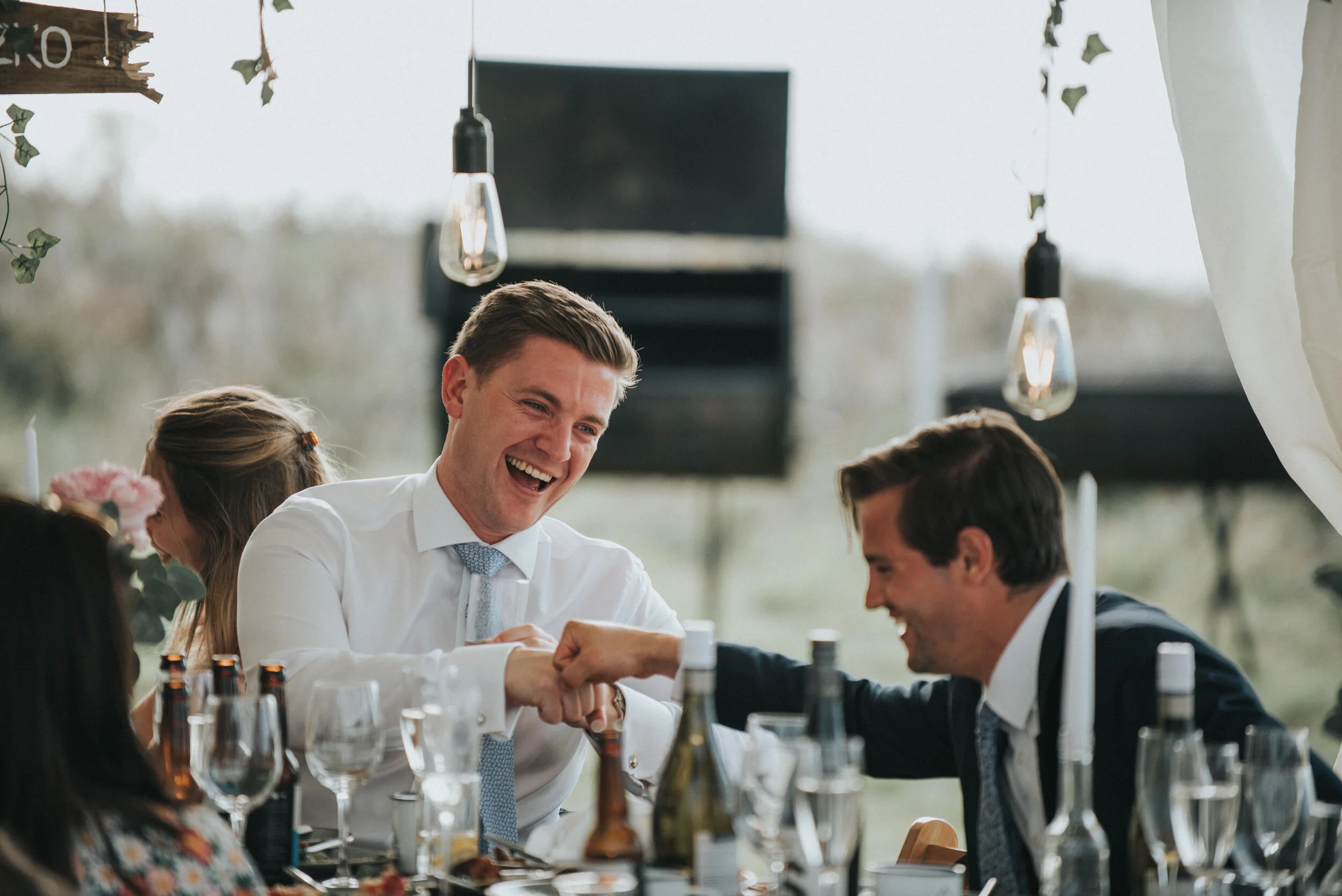 Daymer bay wedding photography in Cornwall captured by Mark Shaw Photography - Two men in formal attire sharing a handshake, smiling, at a decorated event table with other guests, bottles, and glasses, outdoors with hanging light bulbs.