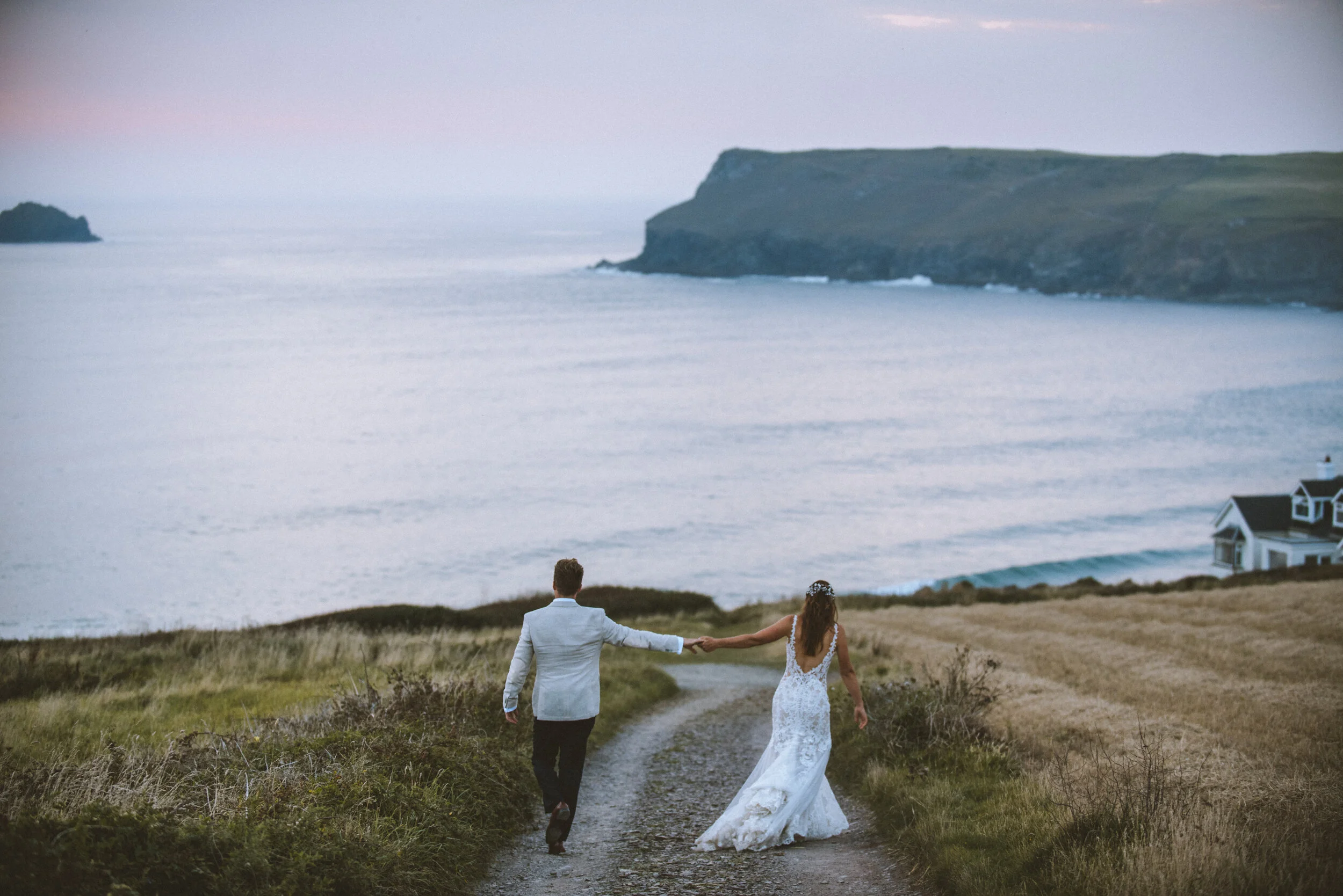 Daymer bay wedding photography in Cornwall captured by Mark Shaw Photography - A bride and groom walking along a dirt path near the coast, holding hands, with the ocean and cliffs in the background at sunset.