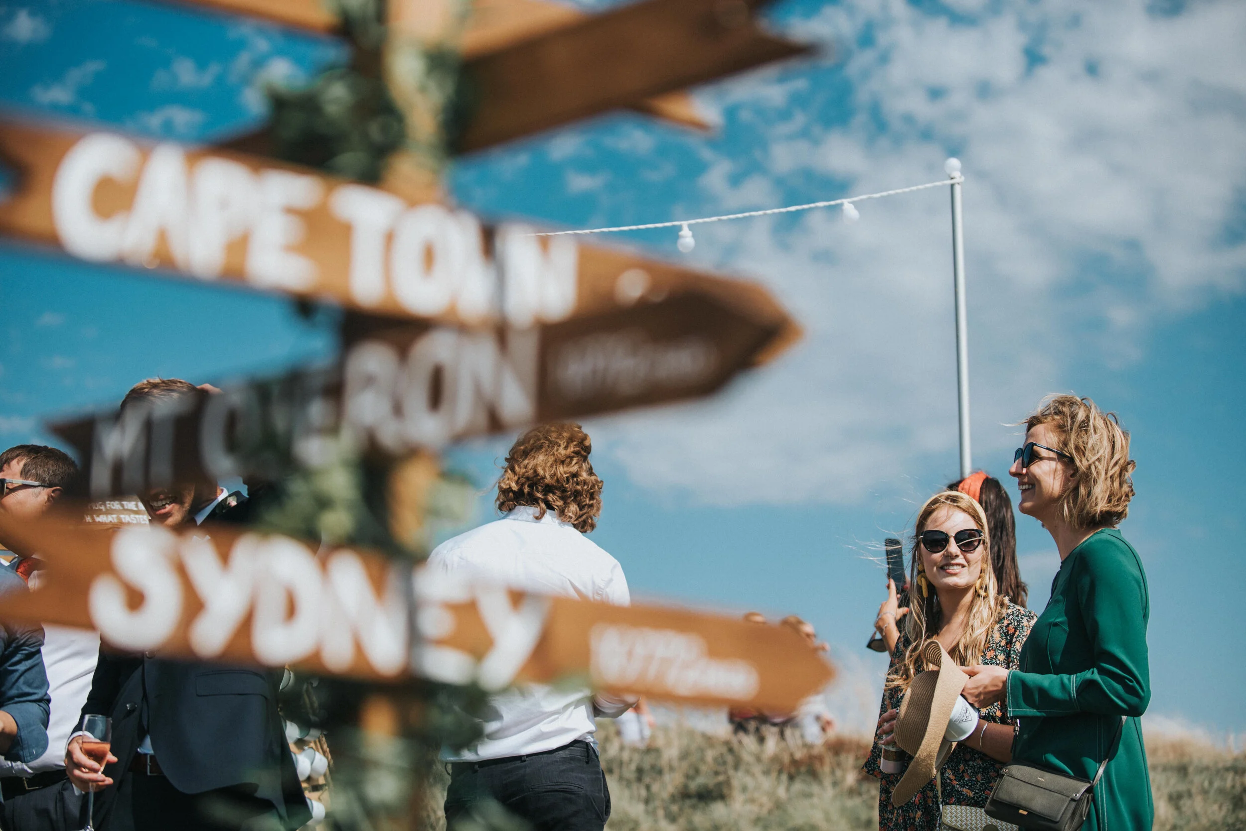 Isles of scilly photography. A group of people socializing outdoors on a sunny day, with a wooden sign in the foreground pointing toward 'CAPE TOWN' and 'SYDNEY'.