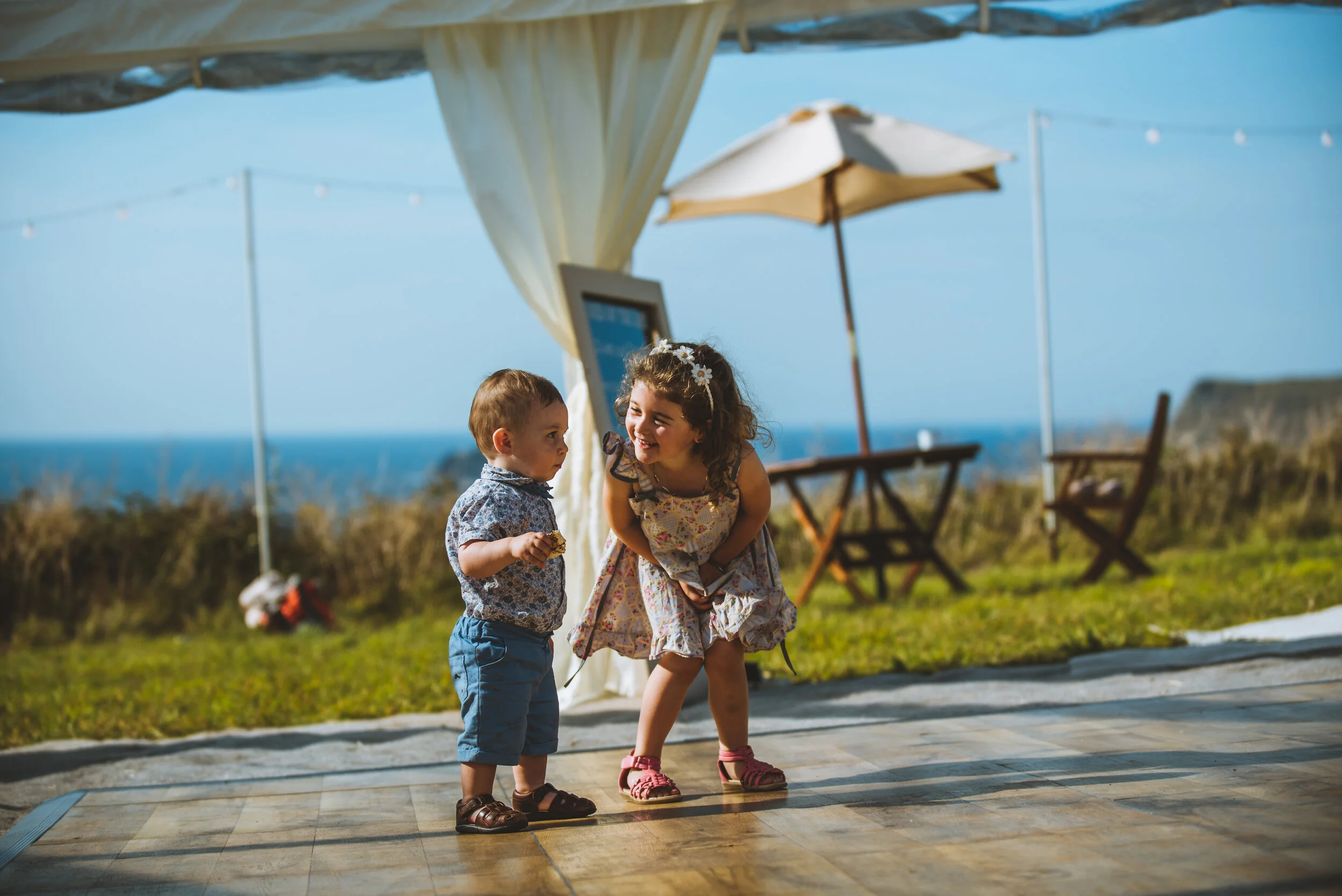 Daymer bay wedding photography in Cornwall captured by Mark Shaw Photography - Two young children, a boy and a girl, outdoors on a sunny day, playing and smiling at each other. The girl is bending slightly toward the boy, who is holding a snack.