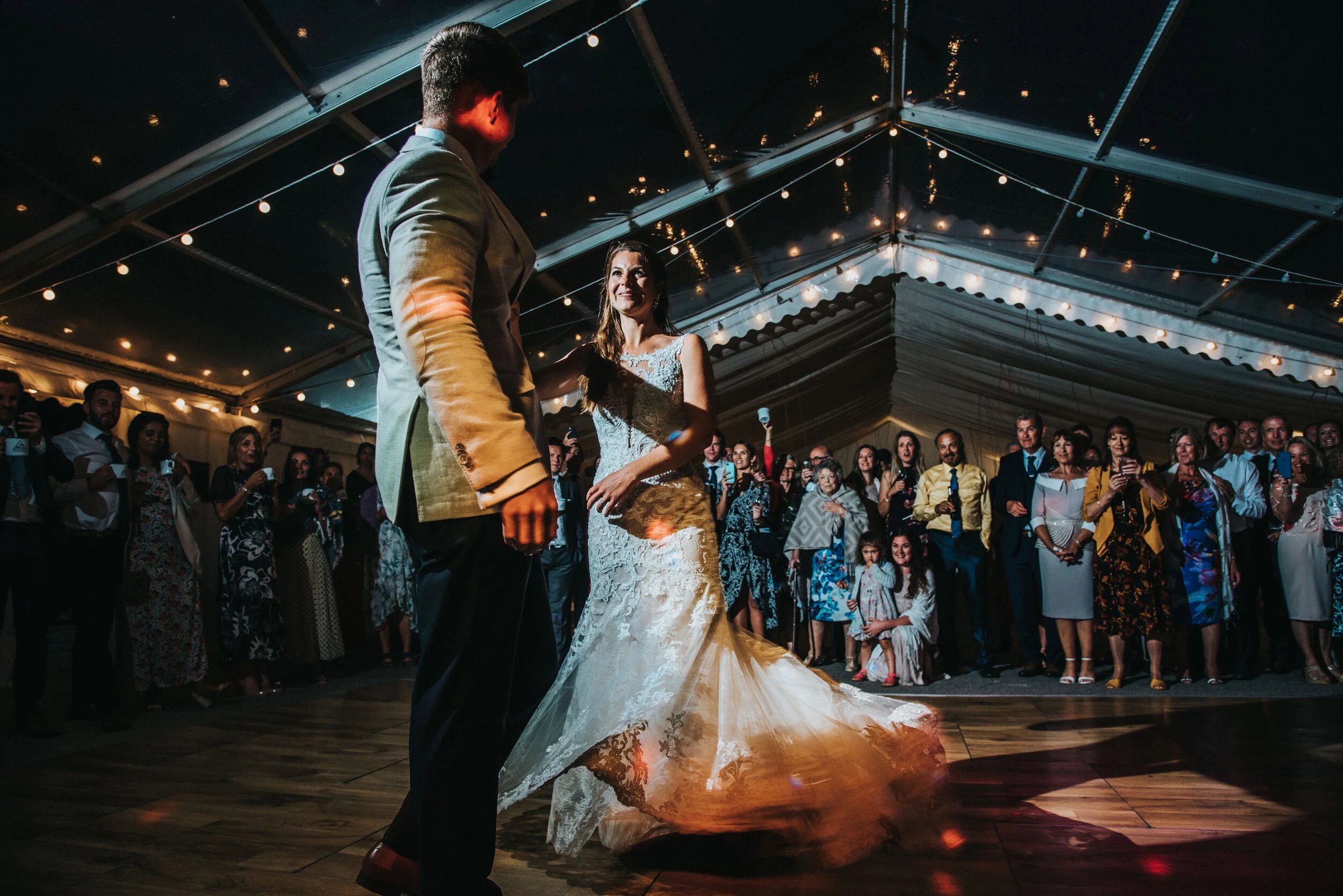 Daymer bay wedding photography captured by Cornwall wedding photographer Mark Shaw Photography - A bride and groom are dancing in a tented wedding reception with guests watching, some taking photos, under string lights.