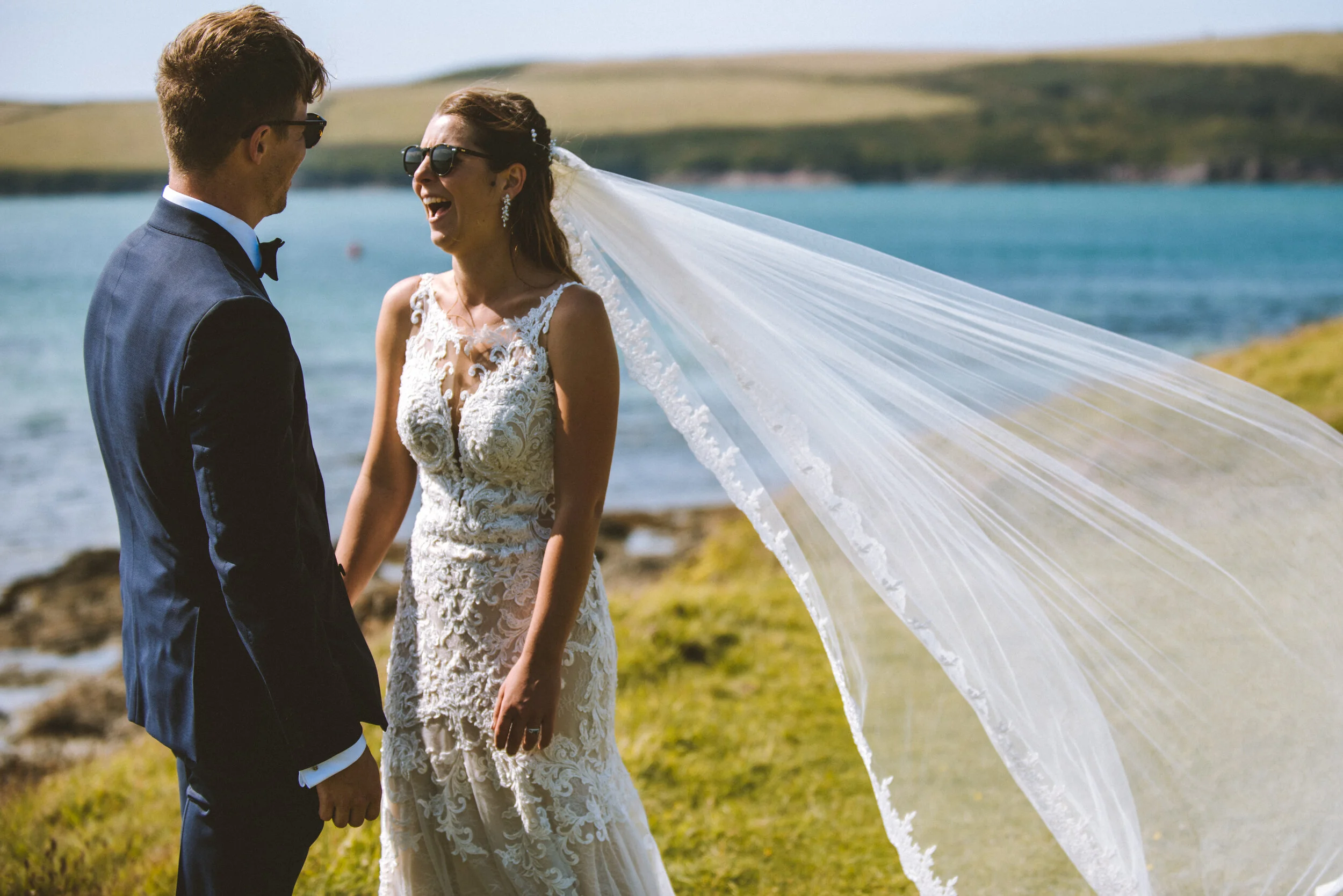 Isles of scilly photography. Bride and groom in wedding attire holding hands and smiling, with a scenic outdoor water and grassy landscape in the background.