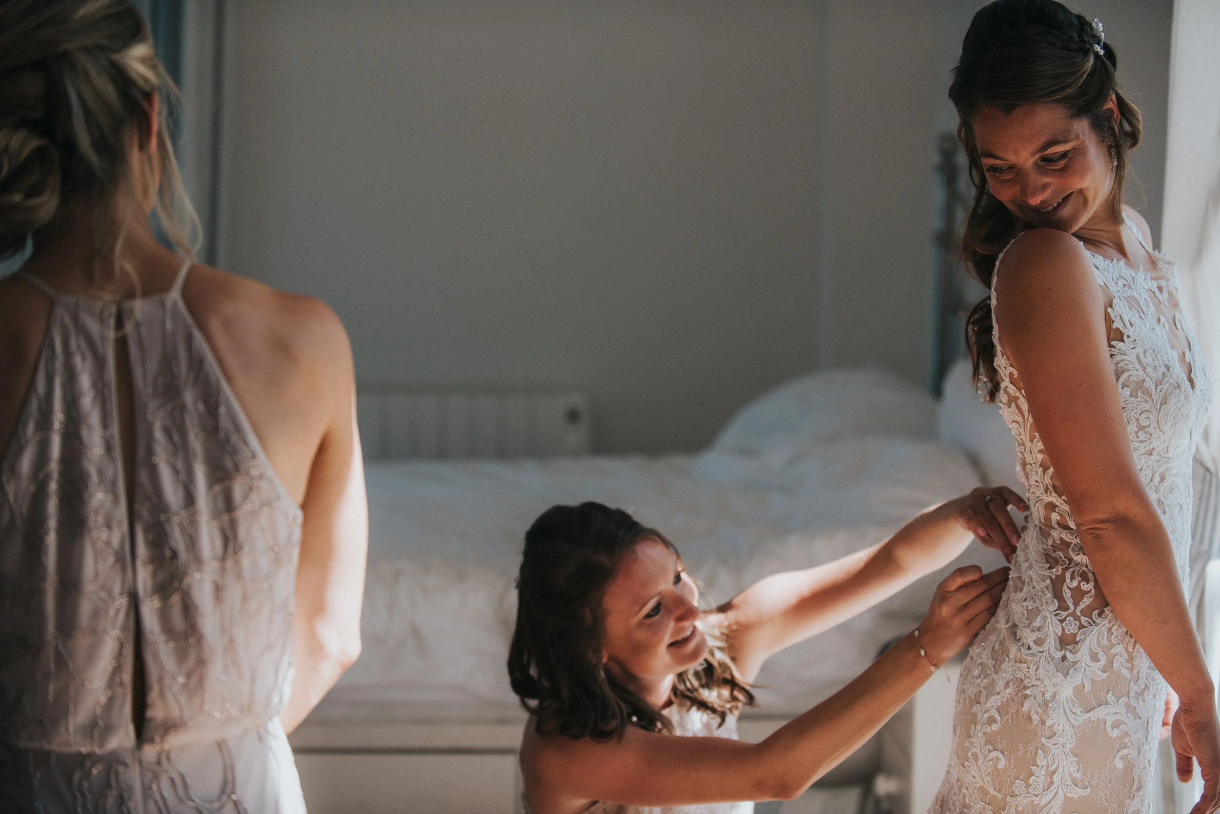 Bridesmaids helping bride get ready, bride in lace dress, bridesmaids in light-colored dresses, interior of a bedroom