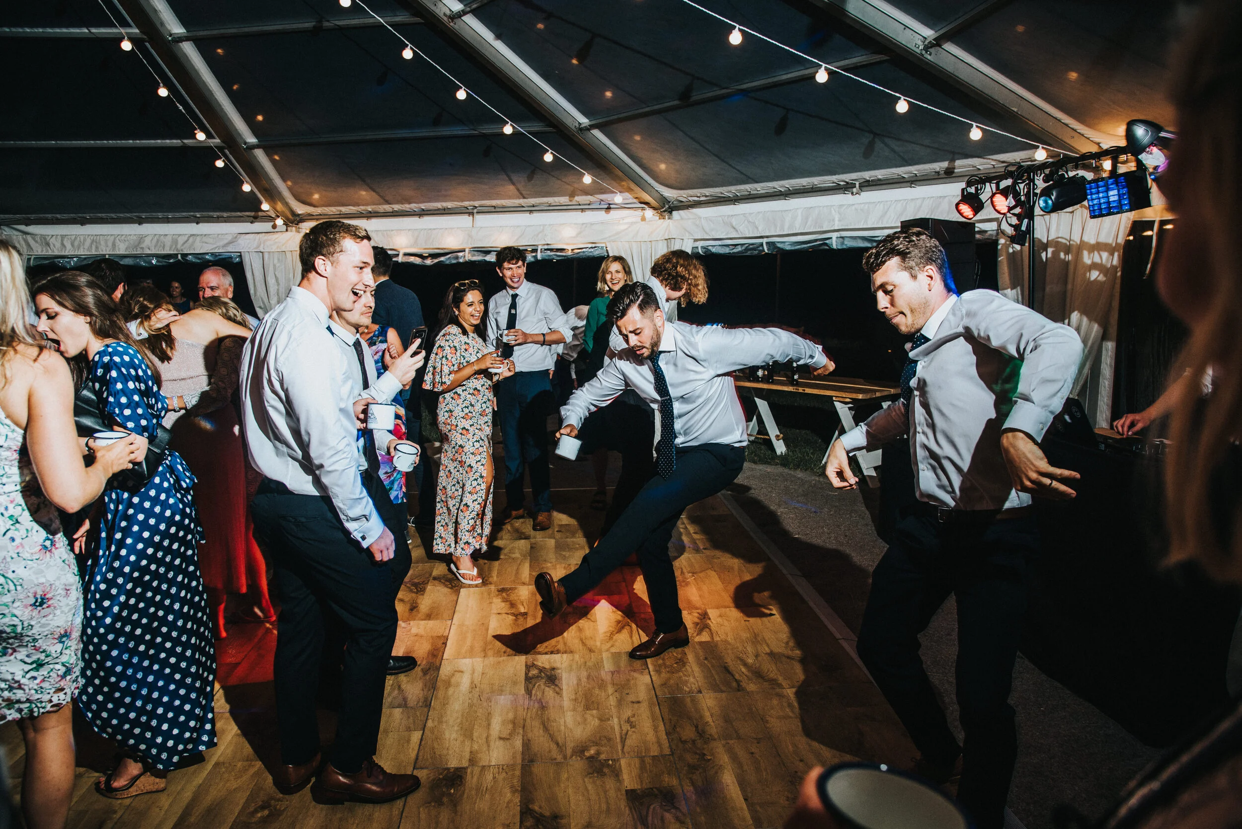 Daymer bay wedding photography captured by Cornwall wedding photographer Mark Shaw Photography -People dancing and enjoying themselves at a nighttime party under a tent with string lights.
