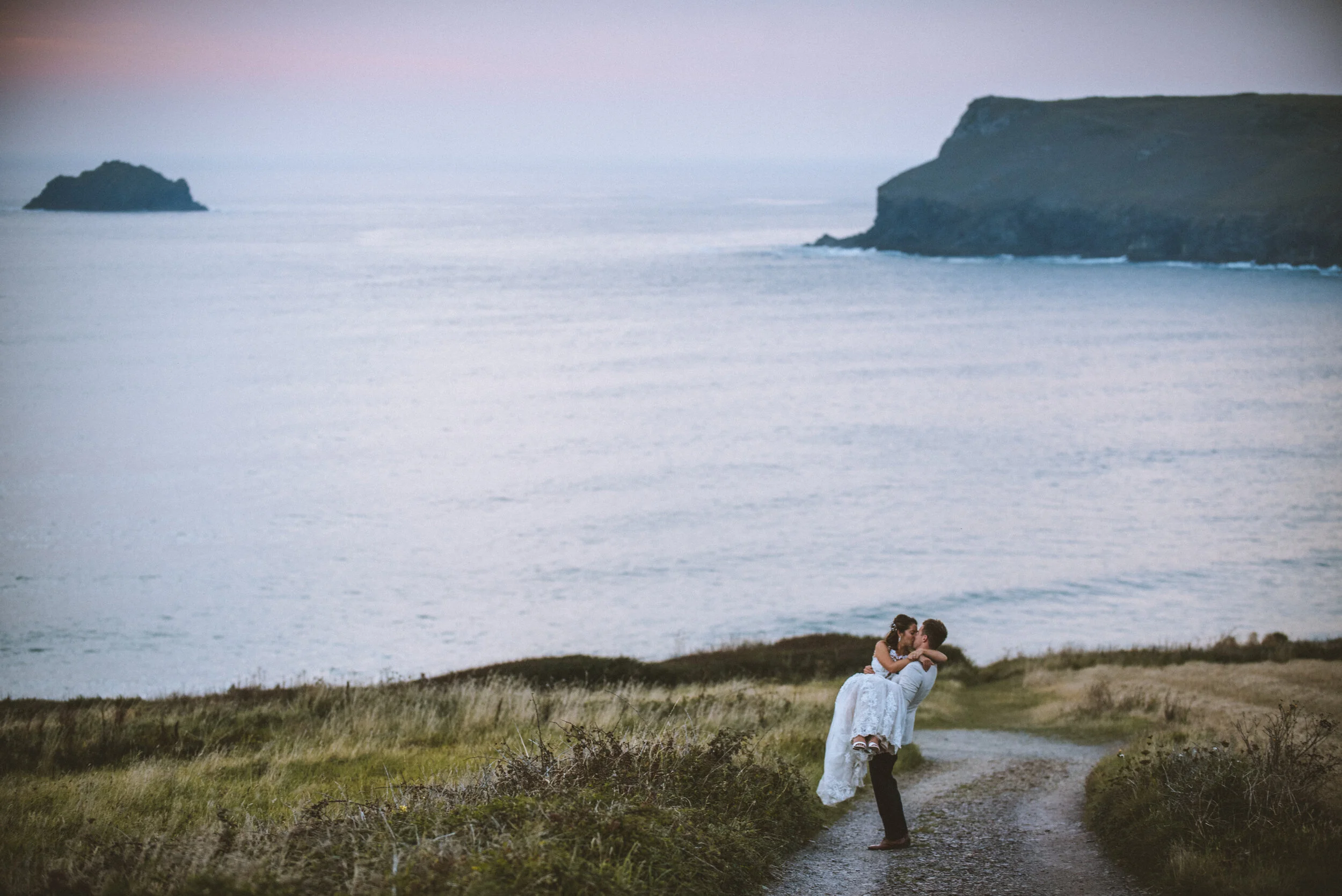 Daymer bay wedding photography in Cornwall captured by Mark Shaw Photography - A couple dressed in wedding attire embracing on a dirt path near the ocean with cliffs in the background at sunset.