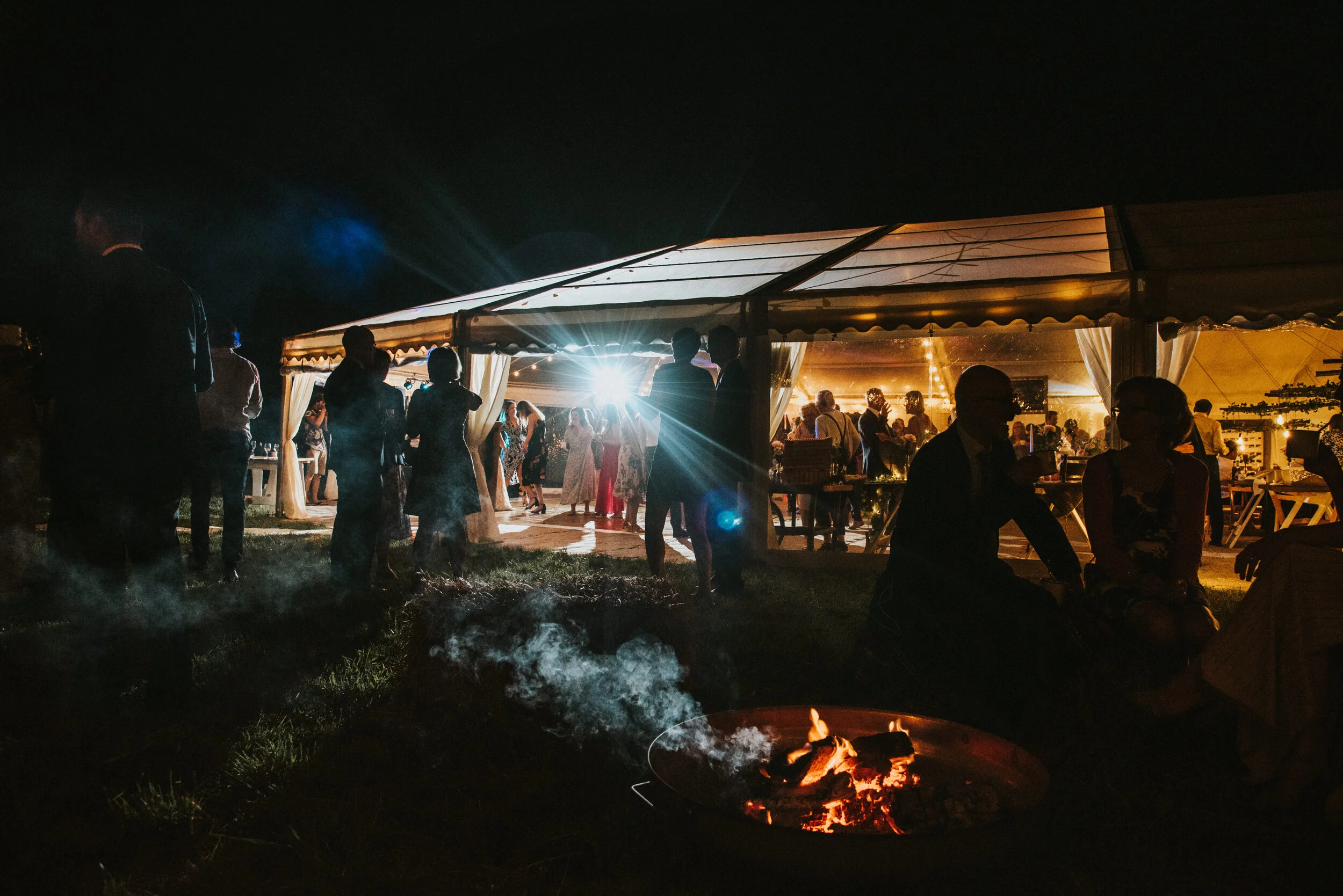 Daymer bay wedding photography captured by Cornwall wedding photographer Mark Shaw Photography - Guests gather and dance at an outdoor nighttime wedding reception under a lit tent, with a firepit in the foreground and string lights in the background.