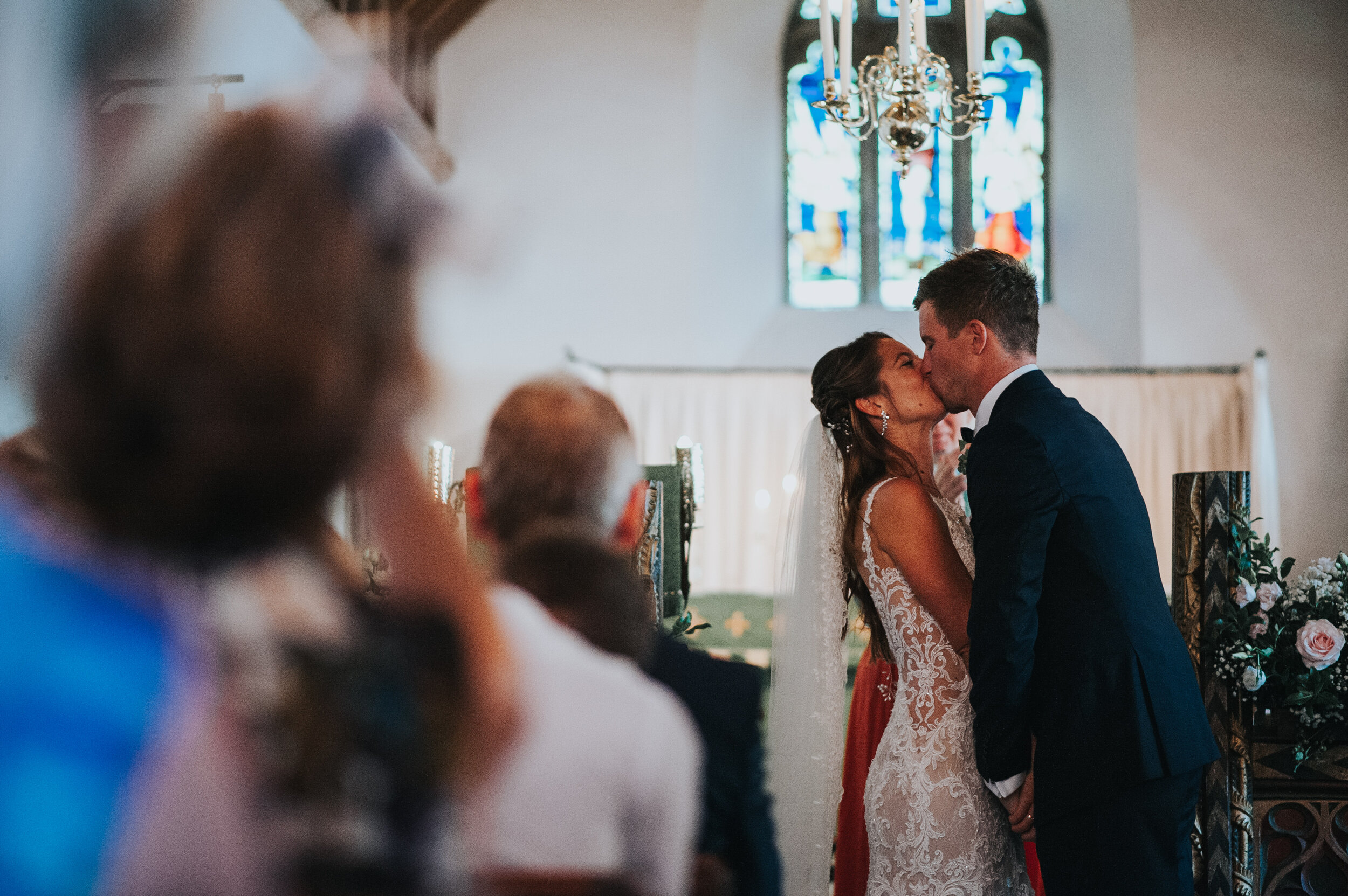 St Enodoc Church wedding photograph, Daymer Bay in Cornwall. A bride and groom kissing during their wedding ceremony inside a church with stained glass windows and a chandelier.