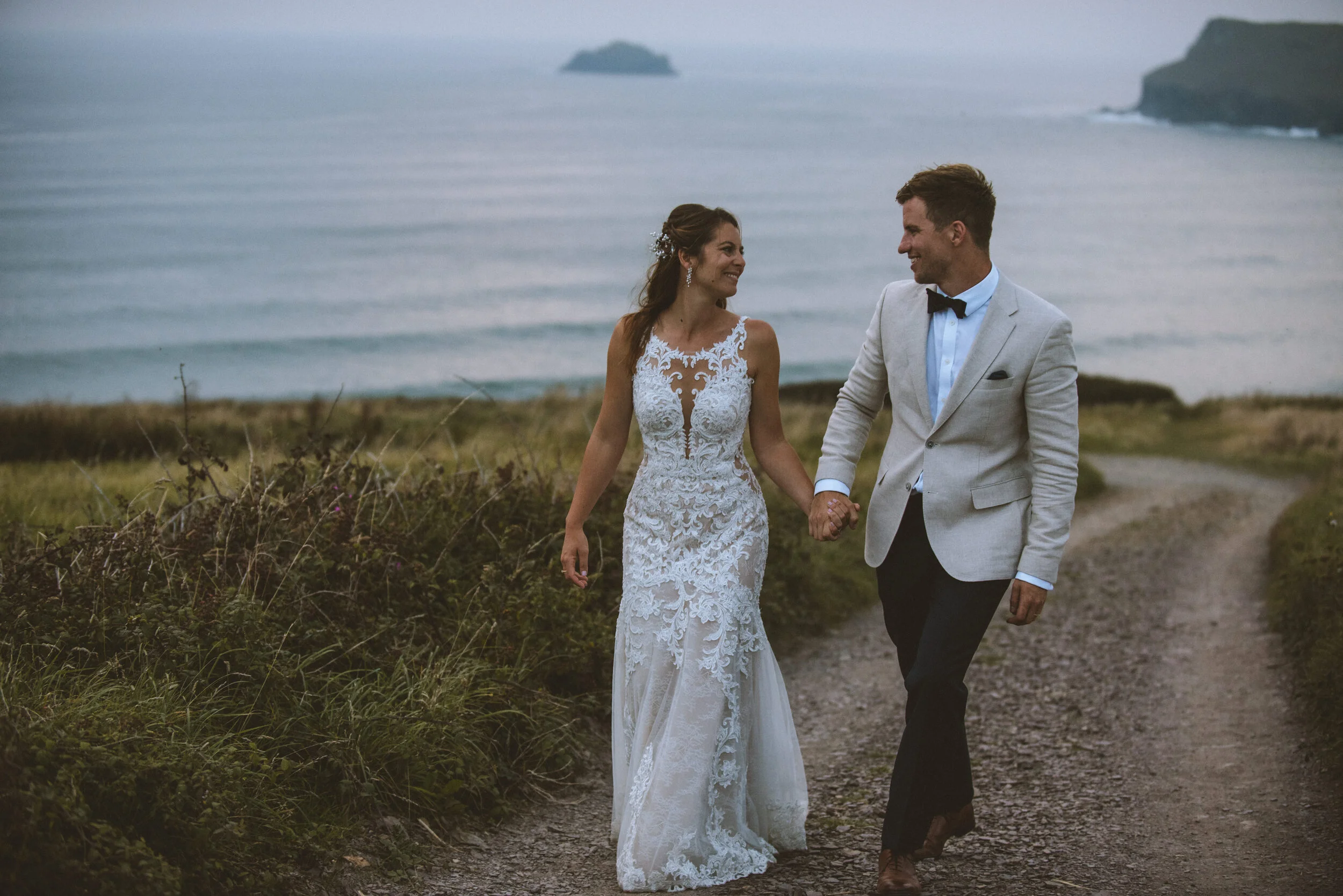 Daymer bay wedding photography captured by Cornwall wedding photographer Mark Shaw Photography - A bride and groom holding hands while walking on a dirt path along the coast, with the ocean and cliffs in the background.