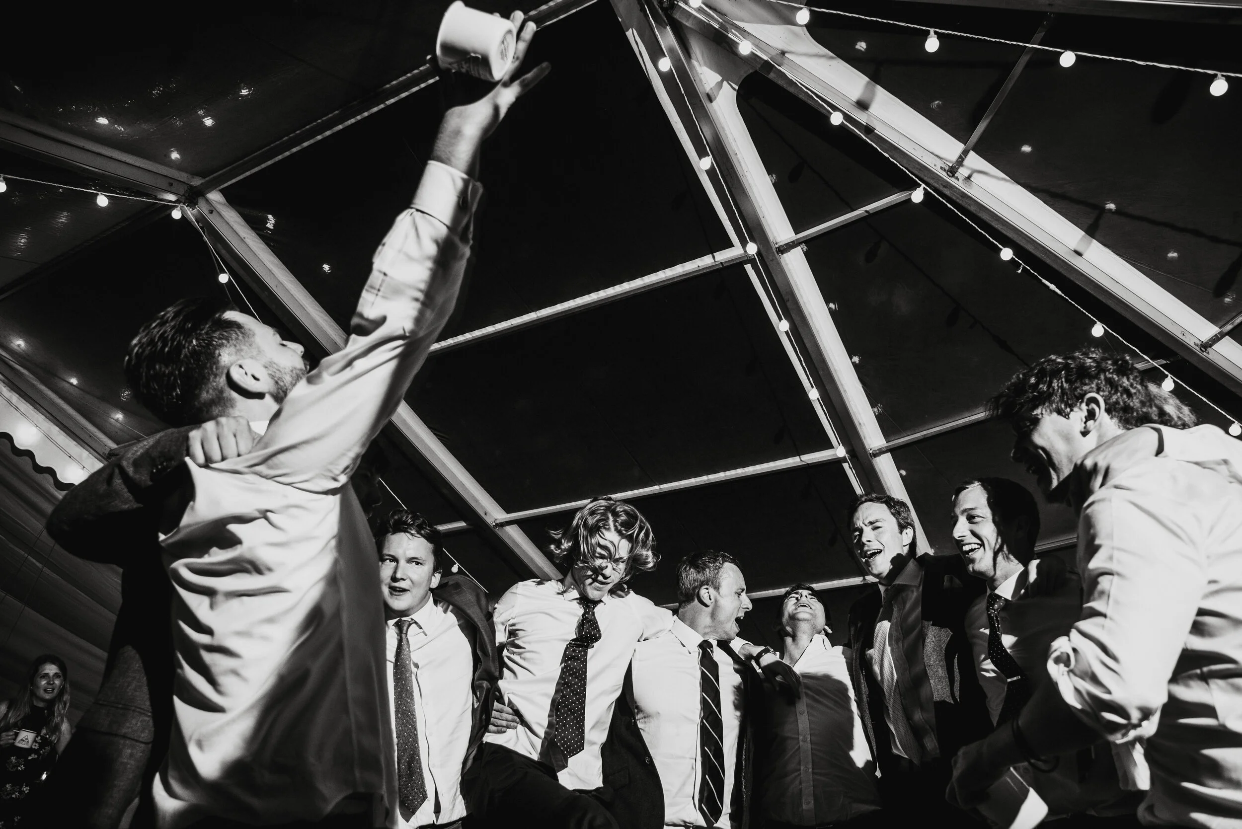 Daymer bay wedding photography captured by Cornwall wedding photographer Mark Shaw Photography - first dance photo. Group of people at a celebration or party, singing and dancing under string lights in black and white
