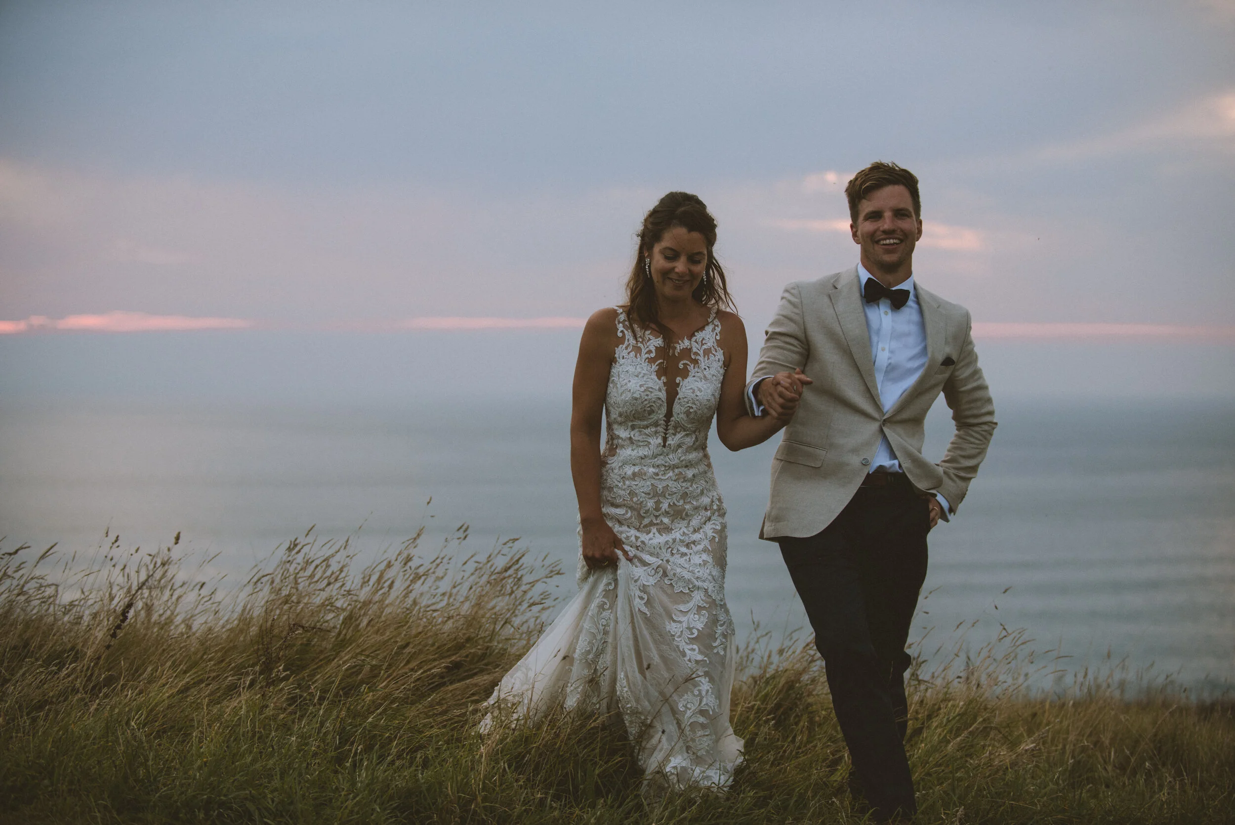 Daymer bay wedding photography captured by Cornwall wedding photographer Mark Shaw Photography - A newlywed couple walking on a grassy field near the ocean at sunset, smiling and holding hands.