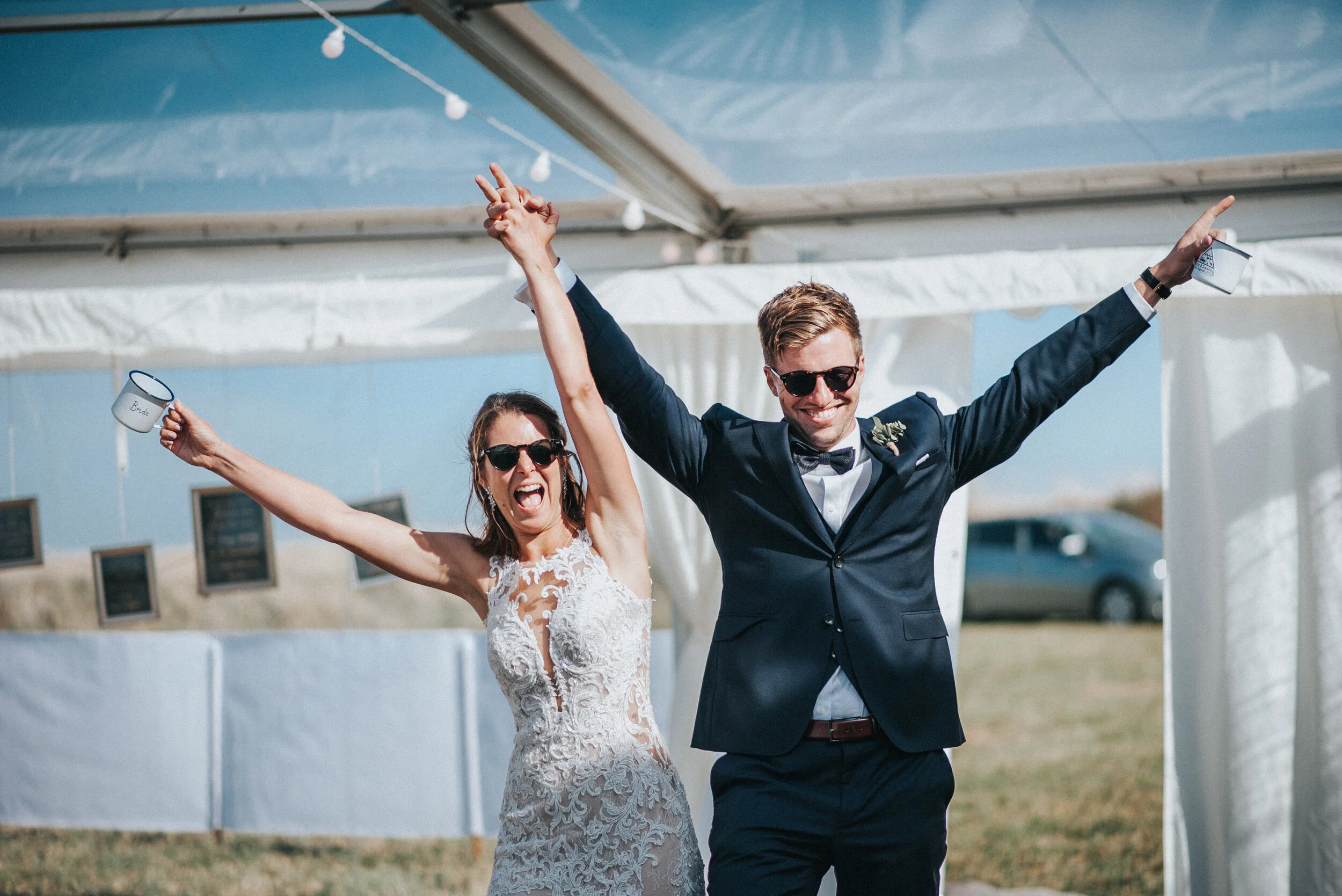Wedding photo at Greenaway Field in Cornwall. A newlywed couple celebrating outdoors, with the bride in a lace wedding dress and the groom in a dark suit, both wearing sunglasses, smiling, and raising their arms in joy under a white tent.