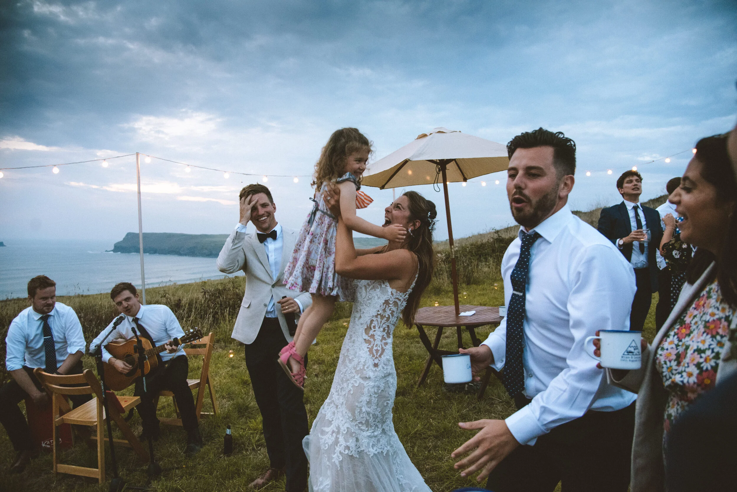 Daymer bay wedding photography captured by Cornwall wedding photographer Mark Shaw Photography - Wedding celebration outdoors with guests dancing, playing guitar, and enjoying the ocean view with string lights and an umbrella.