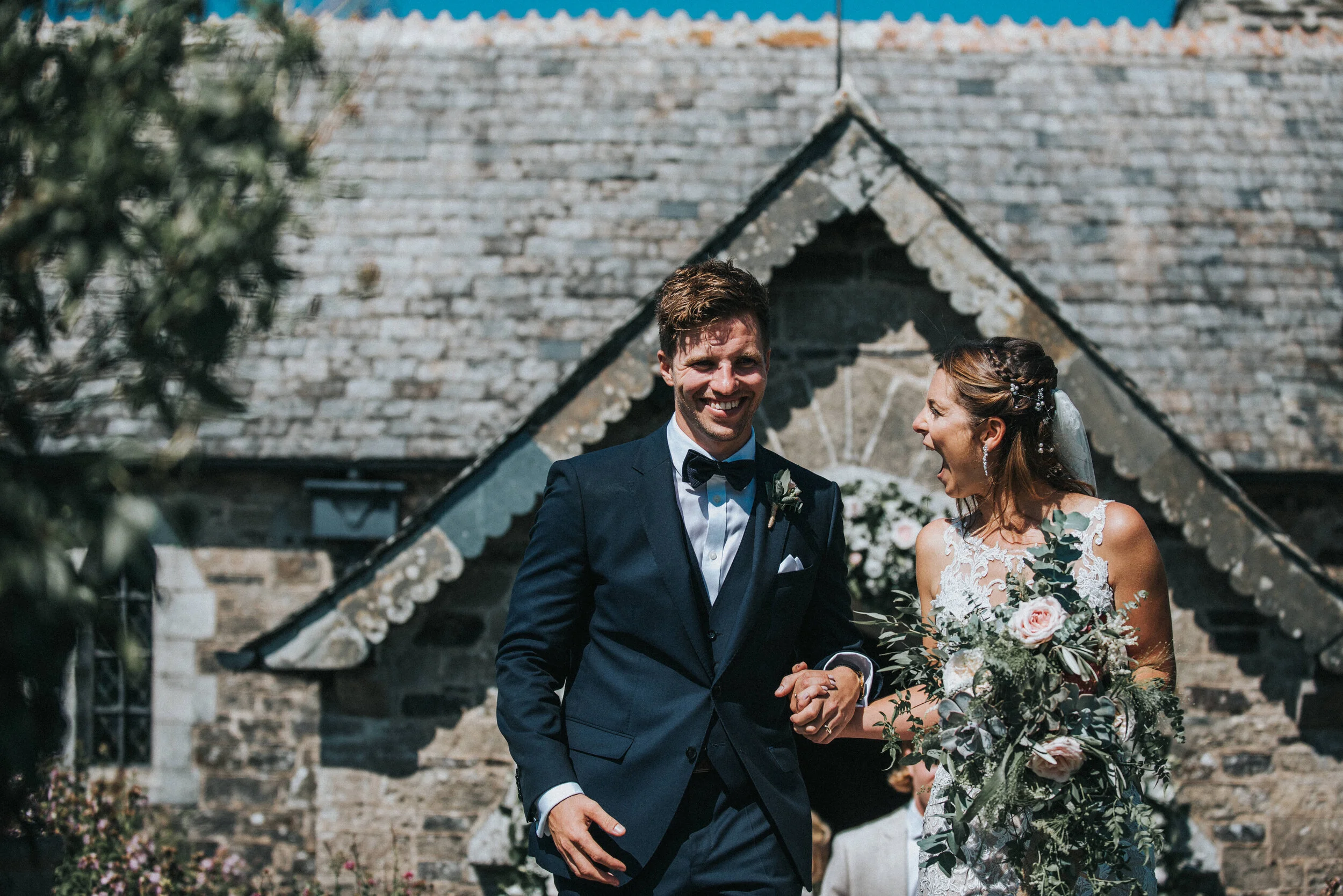 St Enodoc Church wedding photogA happy bride and groom walking arm in arm outside a stone church on their wedding day. The groom is wearing a dark suit and bow tie, and the bride is wearing a lace wedding dress and holding a large bouquet of flowers.