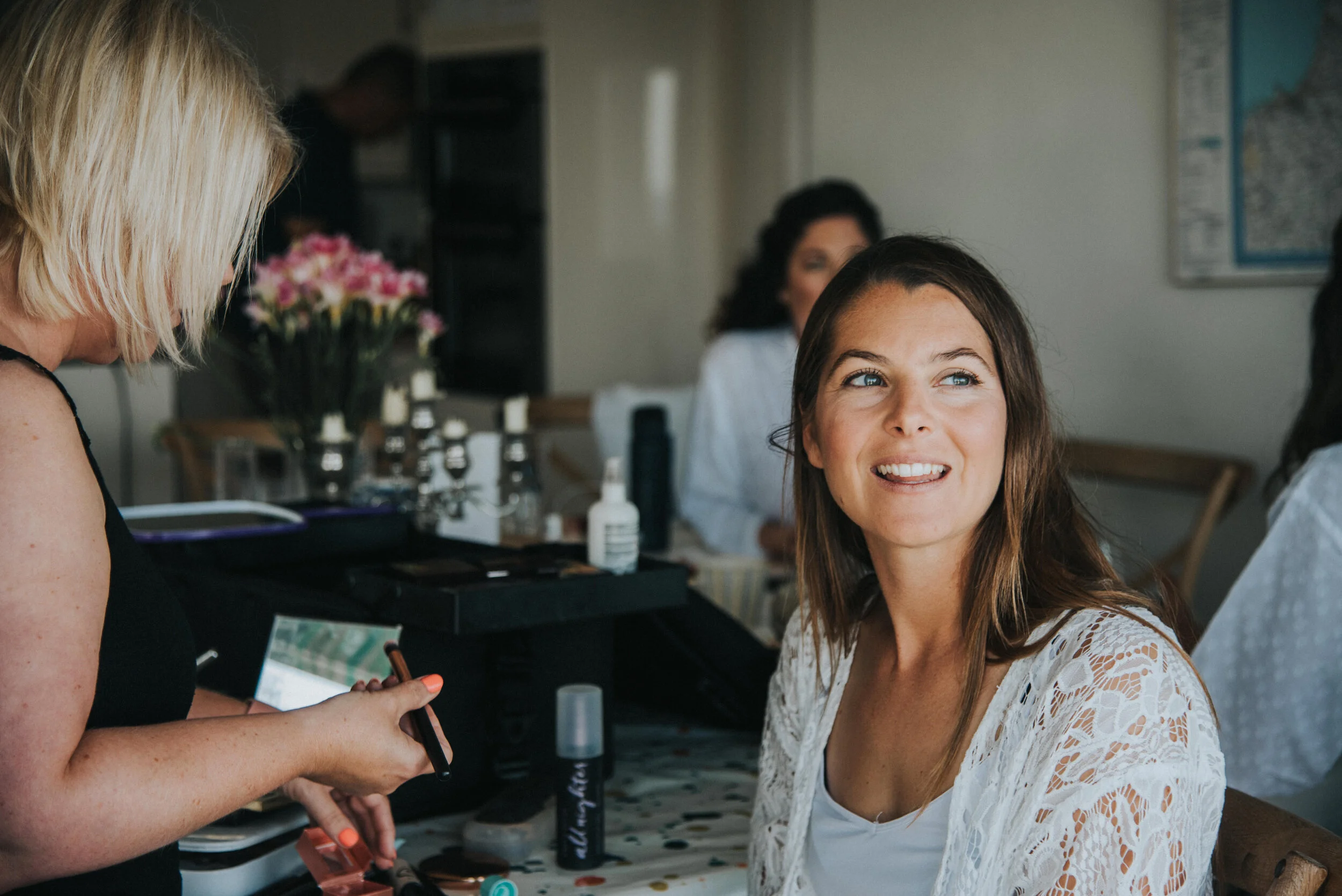 Woman with brown hair smiling and looking to the side at a gathering, with another woman with blonde hair and makeup tools nearby.