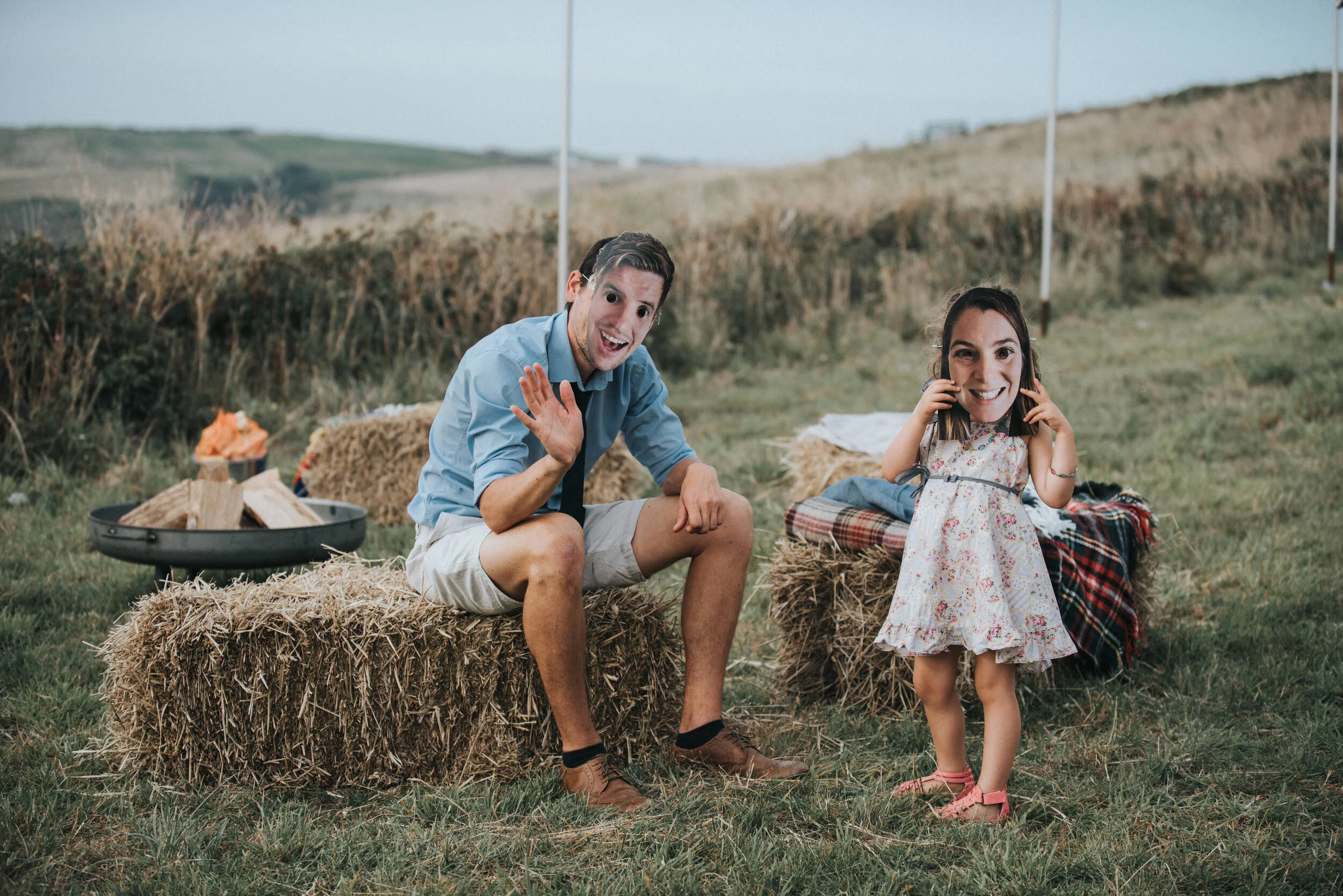 Daymer bay wedding photography in Cornwall captured by Mark Shaw Photography - A man and a young girl sitting on hay bales in an outdoor field, smiling at the camera, with the man waving and the girl talking on a phone, under a partly cloudy sky.