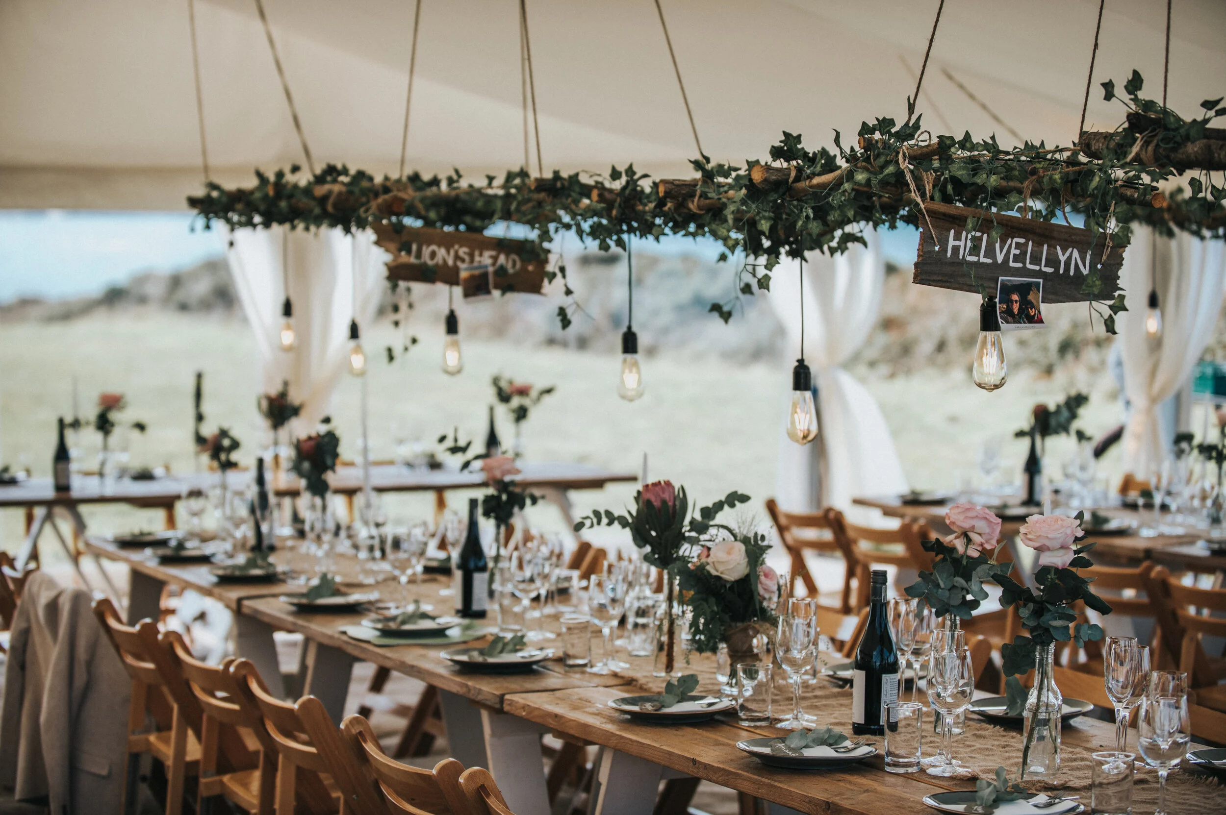 Wedding photo at Greenaway Field in Cornwall. Decorated outdoor wedding reception tent with long wooden tables, floral arrangements, hanging lights, and signs with names 'LION'S HEAD' and 'HELVELLYN.'