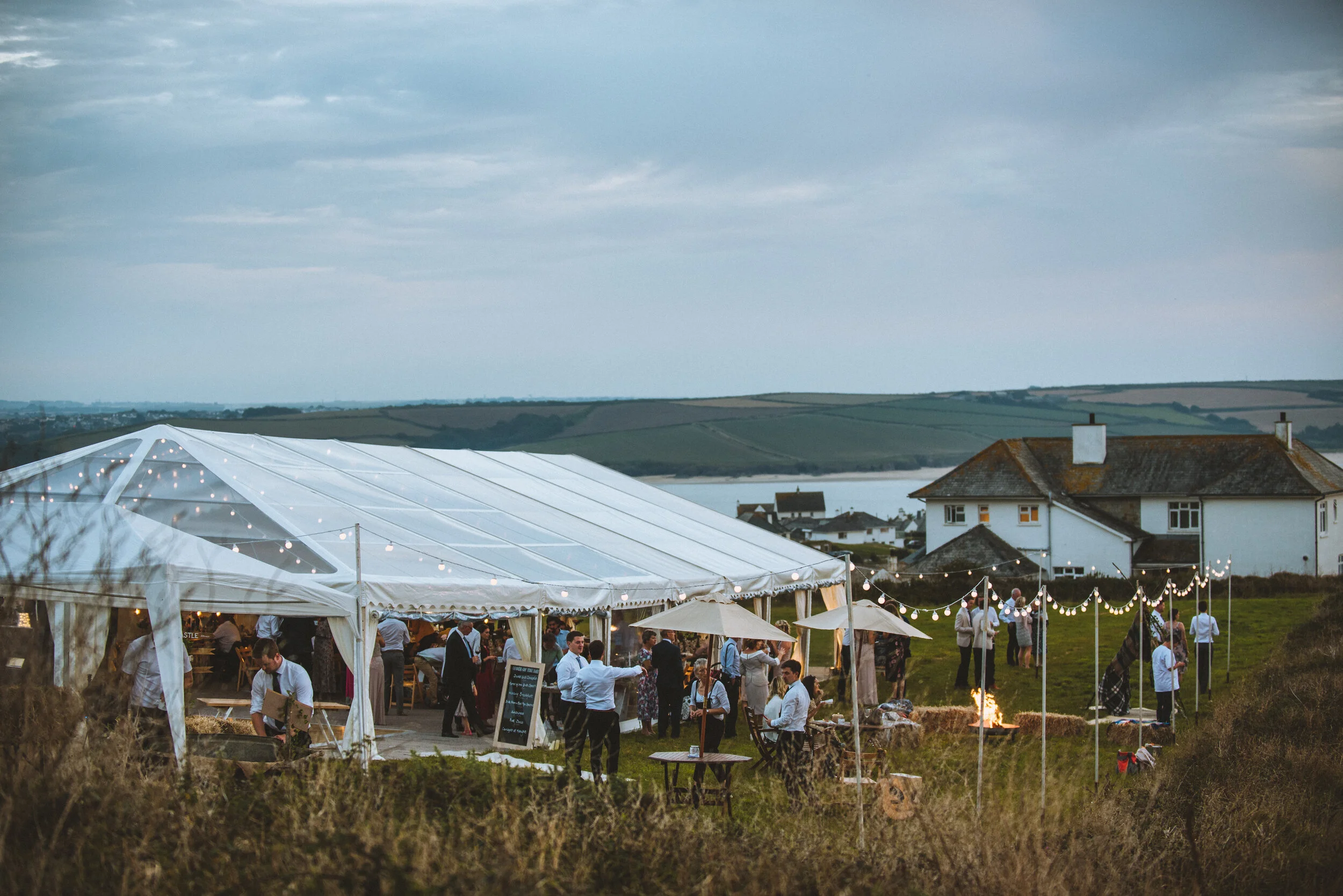 Daymer bay wedding photography in Cornwall captured by Mark Shaw PhotographyOutdoor wedding reception under a large white tent with string lights, tables, and guests socializing near hay bales and a fire pit, with houses and a lake in the background.