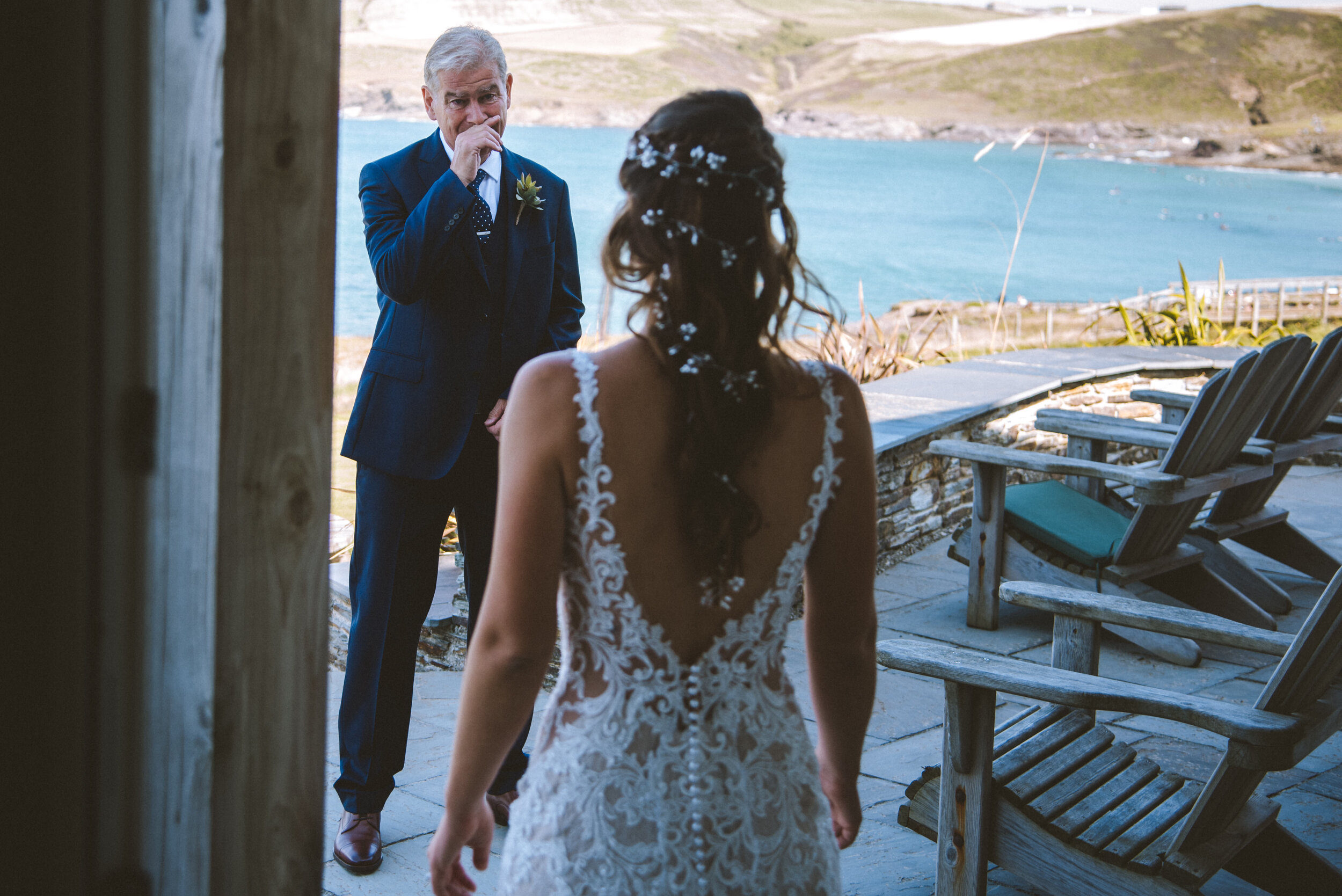 A bride in a lace wedding dress adoringly looks at her father, who is emotional and wiping a tear from his eye, while standing outside near a lake with hills in the background.