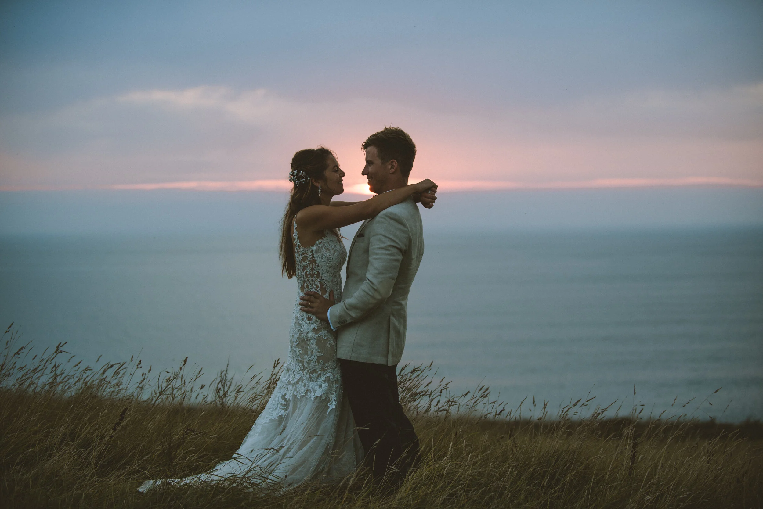 Bride and groom standing closely on a grassy field at sunset, embracing each other.