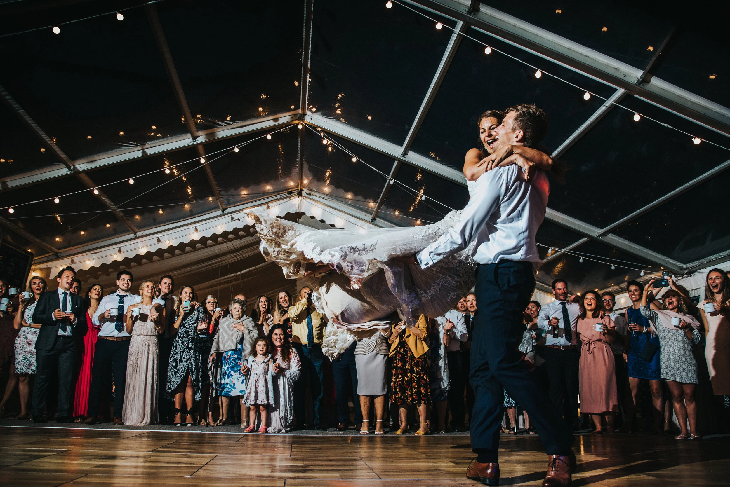 Bride being lifted and spun by groom during a wedding dance under a tent with string lights, surrounded by guests watching and smiling.