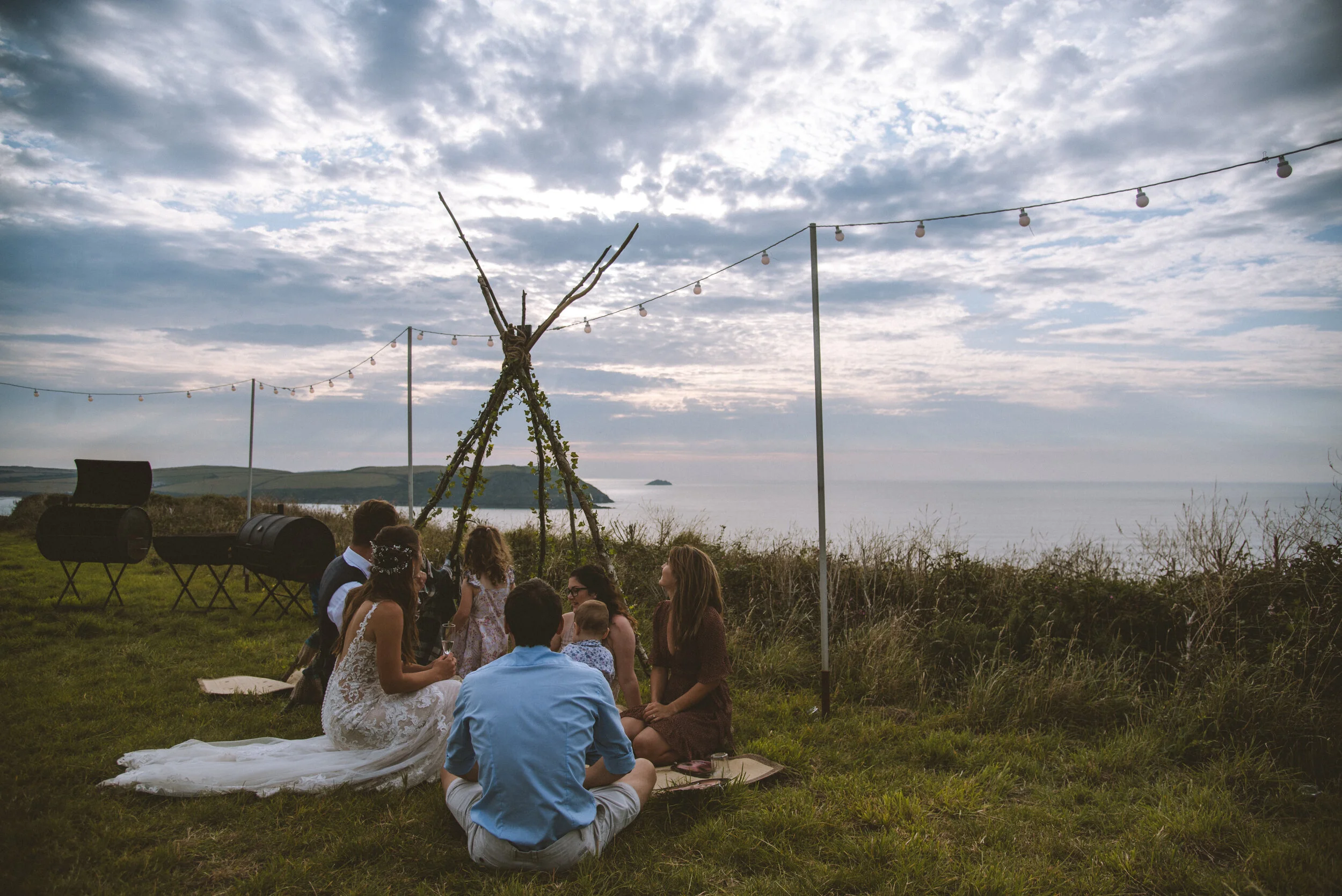 People gathered on grass near the coast, sitting around a small wooden teepee made of sticks, with string lights hanging above, overlooking the ocean and cloudy sky.