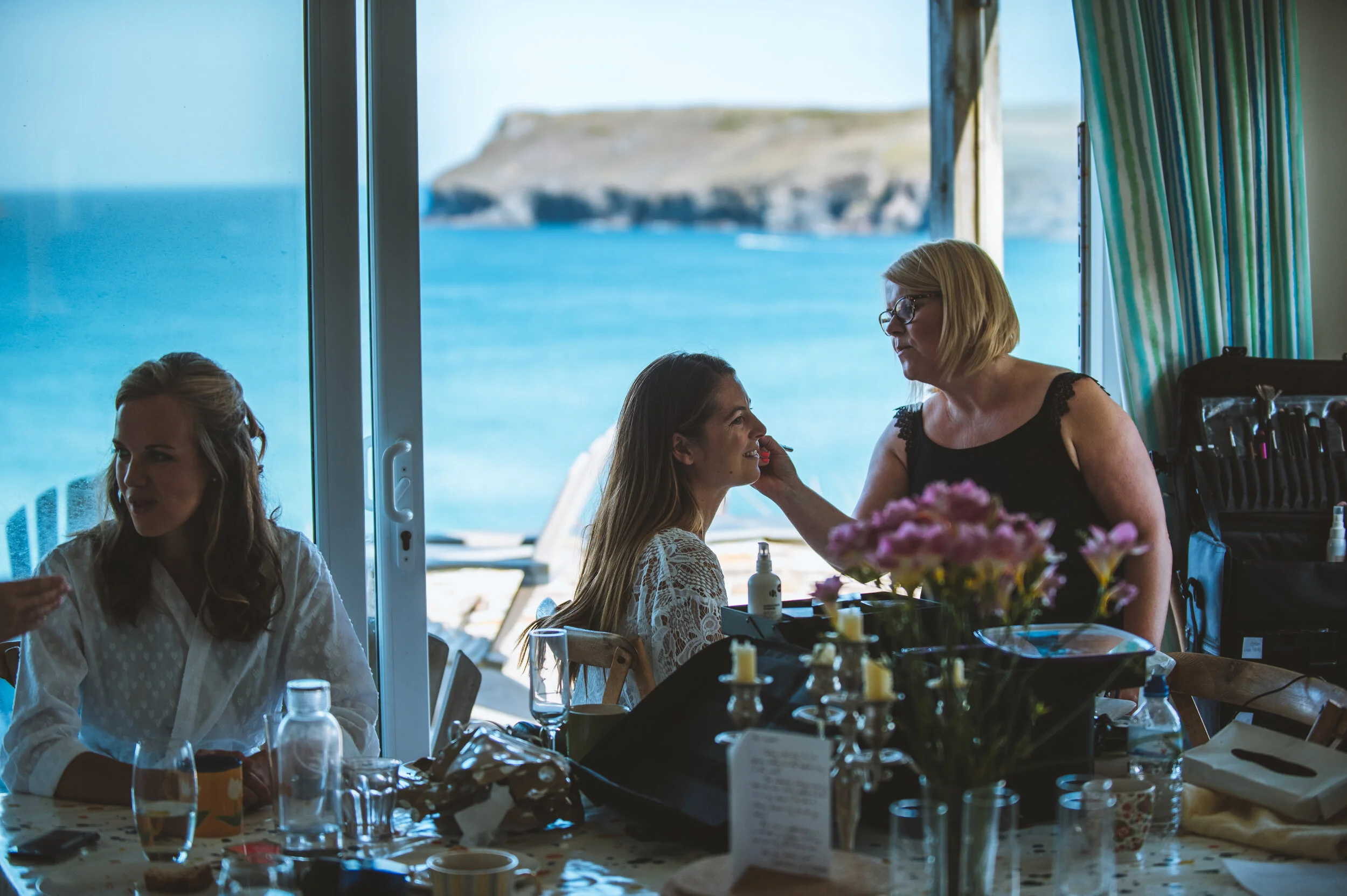 Women getting ready for a special occasion, with a seaside view in the background. One woman is applying makeup to another woman at a table decorated with flowers and candles.