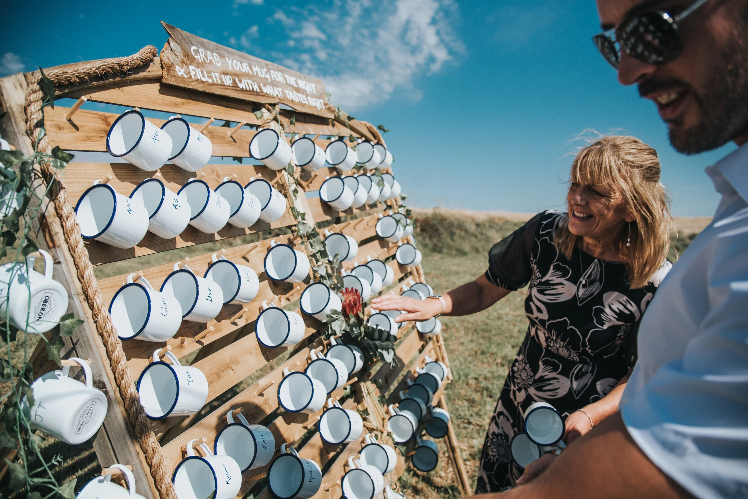 Isles of scilly photography. People at an outdoor wedding activity filling mugs with tea from a display of mugs hanging on a wooden board with a sign that says, "Grab your mug for the night & fill it up with what tastes right."