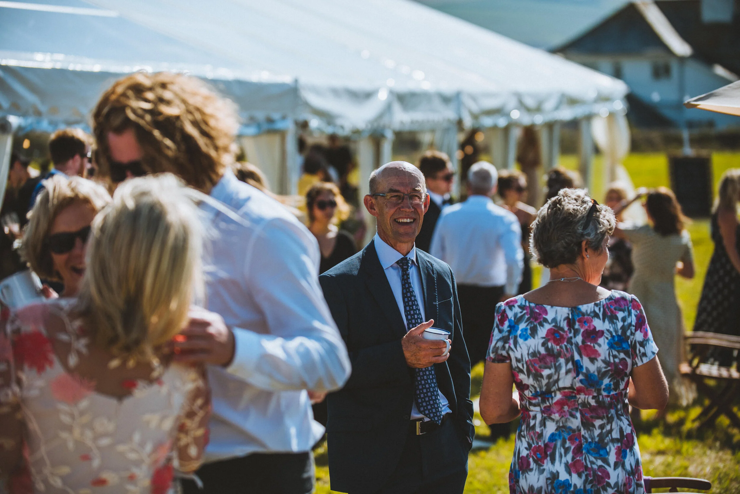 Wedding photo at Greenaway Field in Cornwall. People socializing outdoors at a sunny event with white tents in the background. A man in a dark suit and glasses is smiling, holding a cup. Several women and men are chatting and enjoying the gathering.