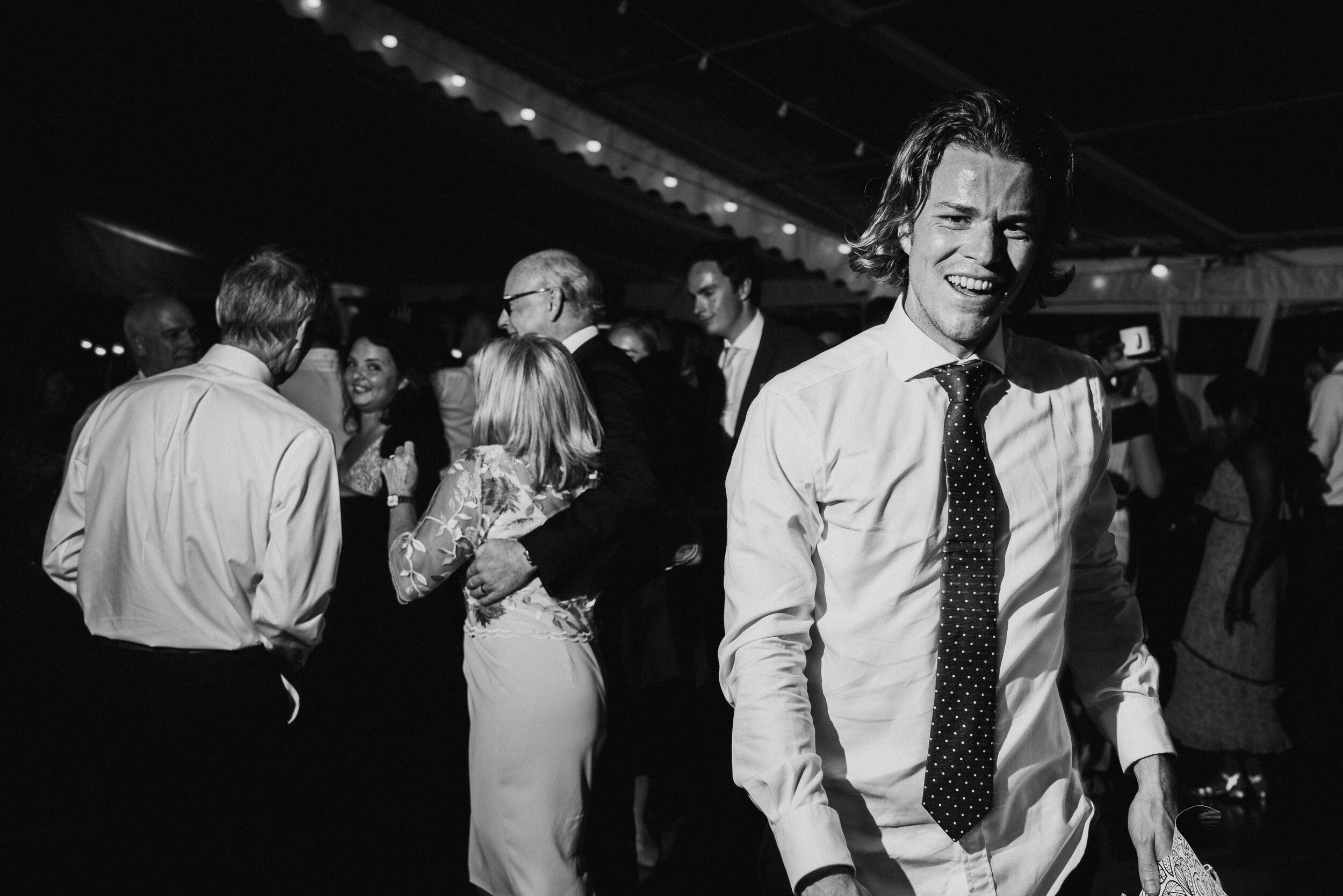 Daymer bay wedding photography captured by Cornwall wedding photographer MarA young man with shoulder-length hair, wearing a white shirt and striped tie, smiling at a party or celebration with other people in the background, some dancing and talking.