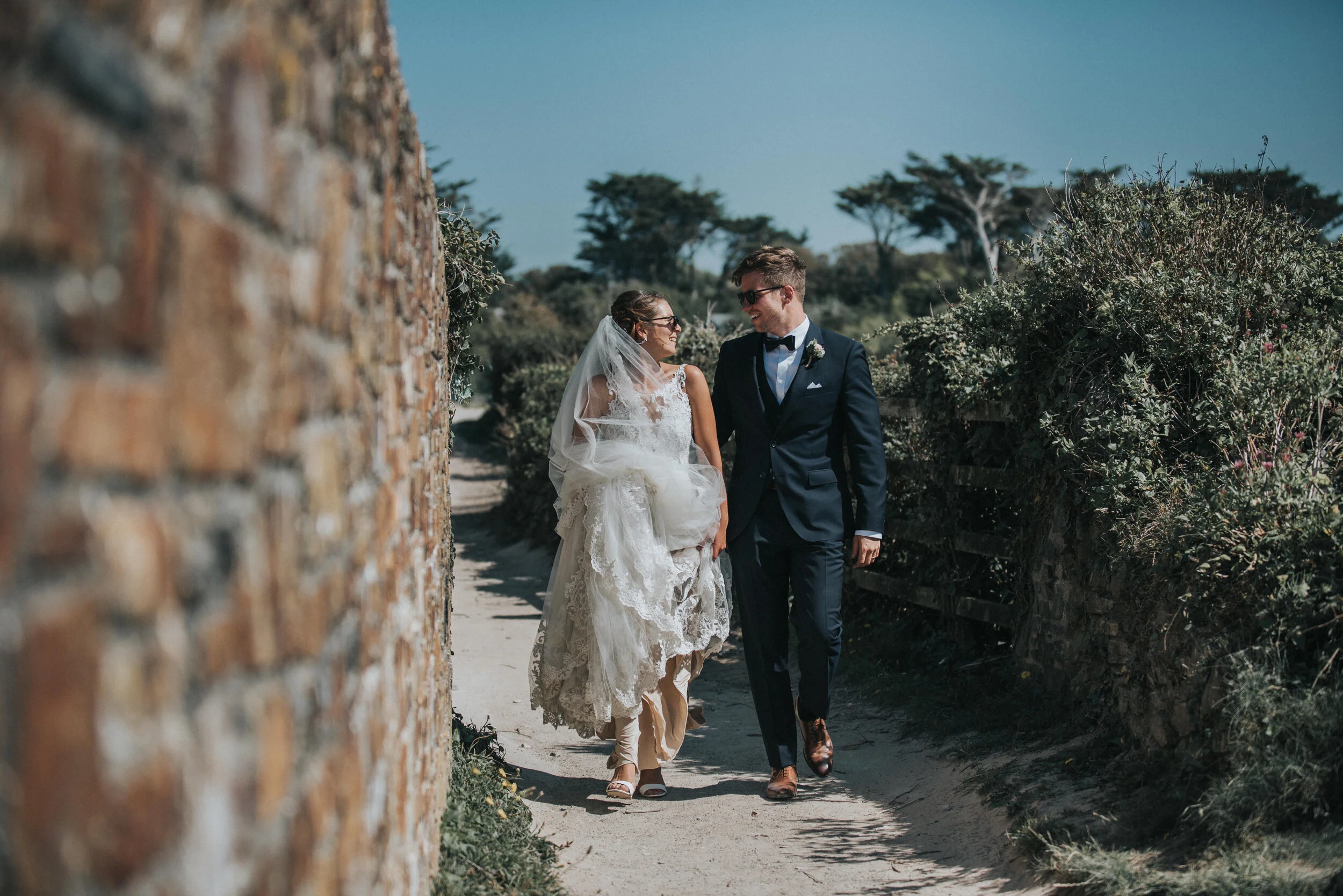 Isles of scilly photography. Bride and groom in wedding attire walking joyfully on a narrow dirt path, surrounded by greenery and stone walls on a sunny day.