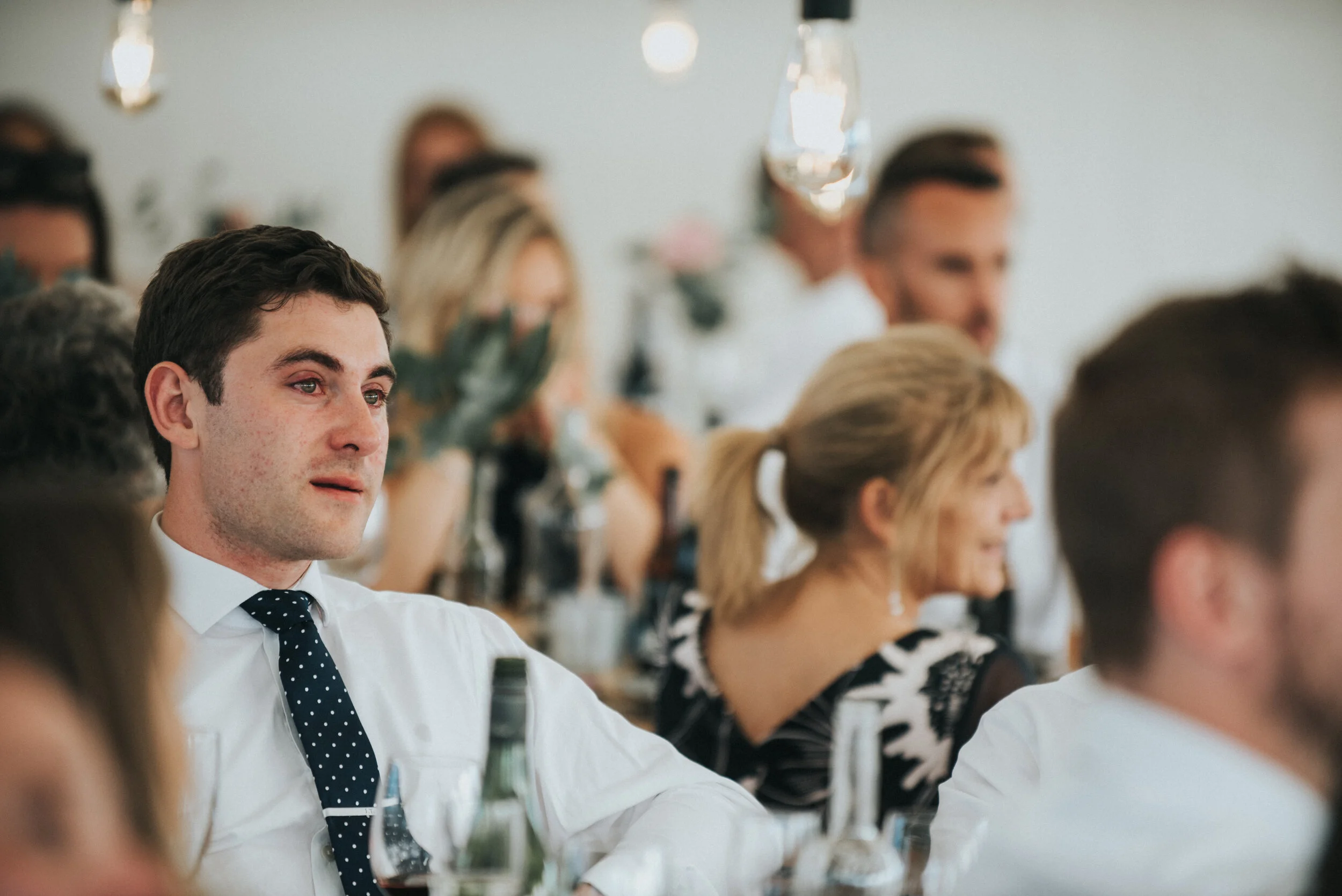 Daymer bay wedding photography in Cornwall captured by Mark Shaw Photography - A young man with dark hair wearing a white shirt and black polka dot tie sitting at a table during a formal event, with other guests blurred in the background.