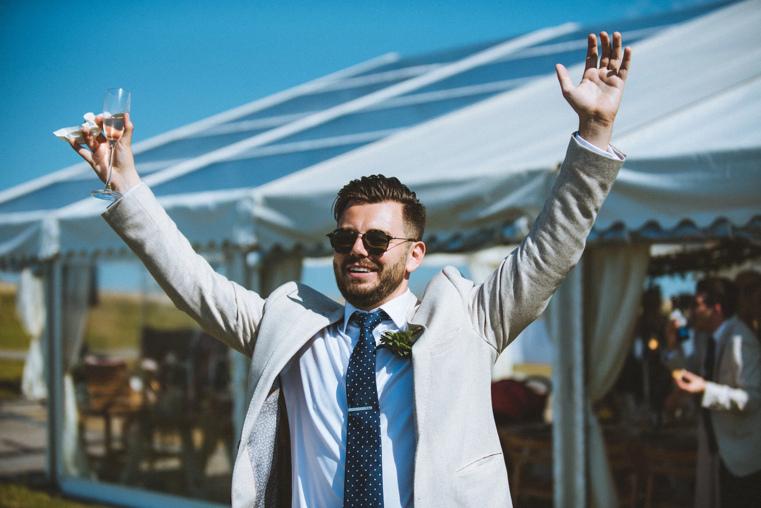 Wedding photo at Greenaway Field in Cornwall. A man in a light-colored suit, sunglasses, and a tie holding a glass of wine or champagne, smiling with arms raised at an outdoor event with a tent and other people in the background.