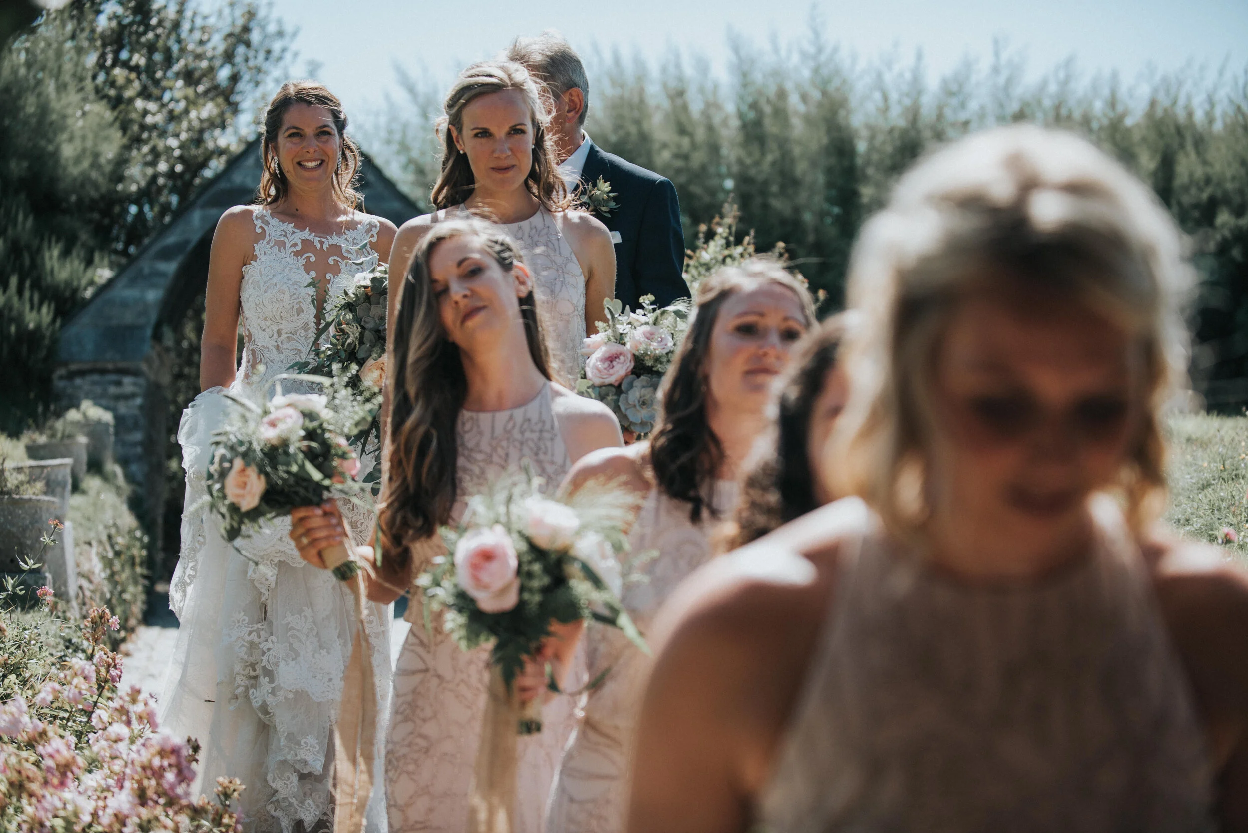 Daymer bay wedding photography in Cornwall captured by Mark Shaw Photography - Wedding party walking outdoors with bridesmaids holding bouquets, bride in white gown at back, sunlit garden setting with trees and flowers.