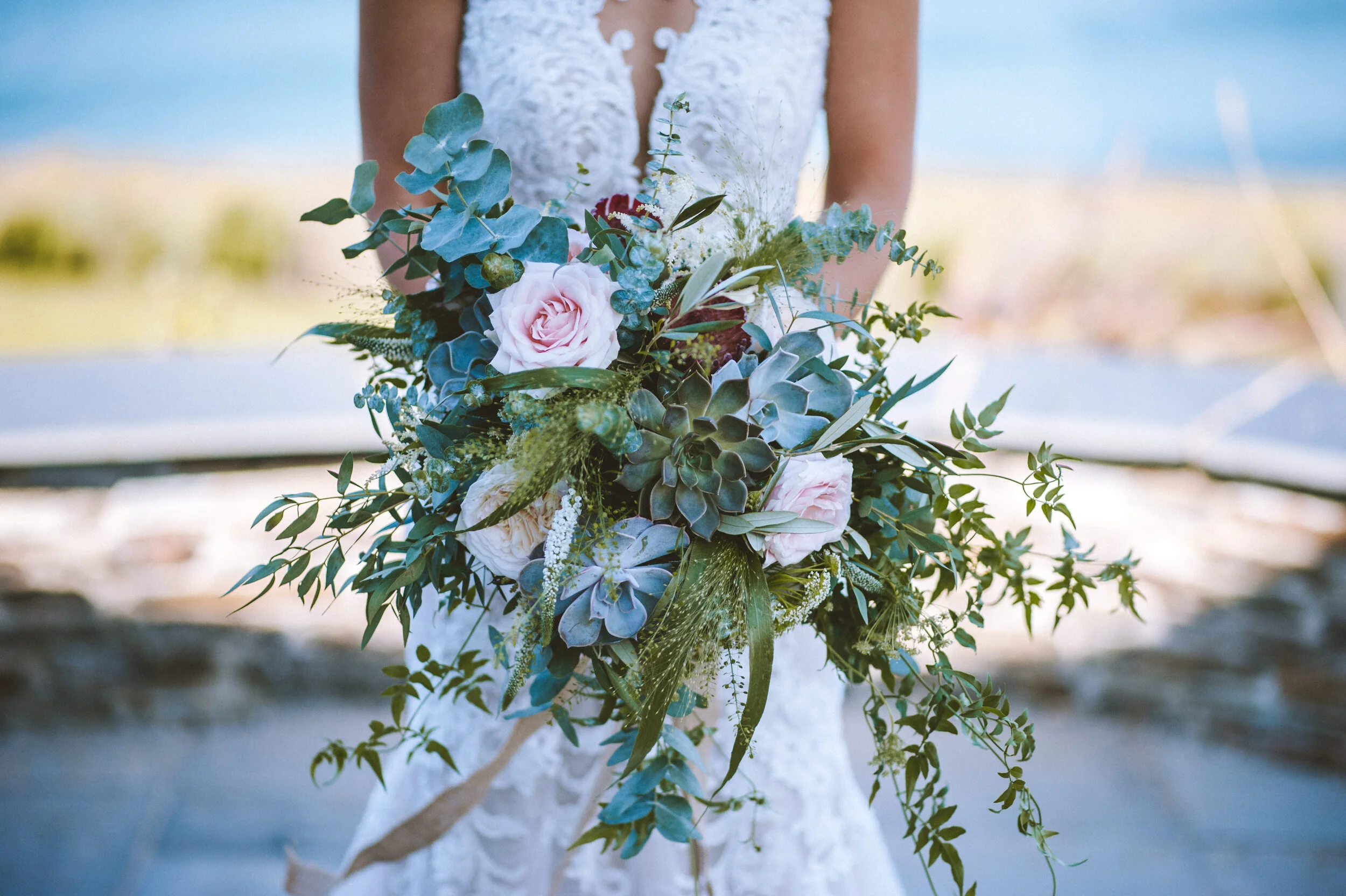 Daymer bay wedding photography in Cornwall captured by Mark Shaw Photography - A person in a white lace wedding dress holds a large bouquet of pink roses, green succulents, eucalyptus, and other greenery against a blurred outdoor background.