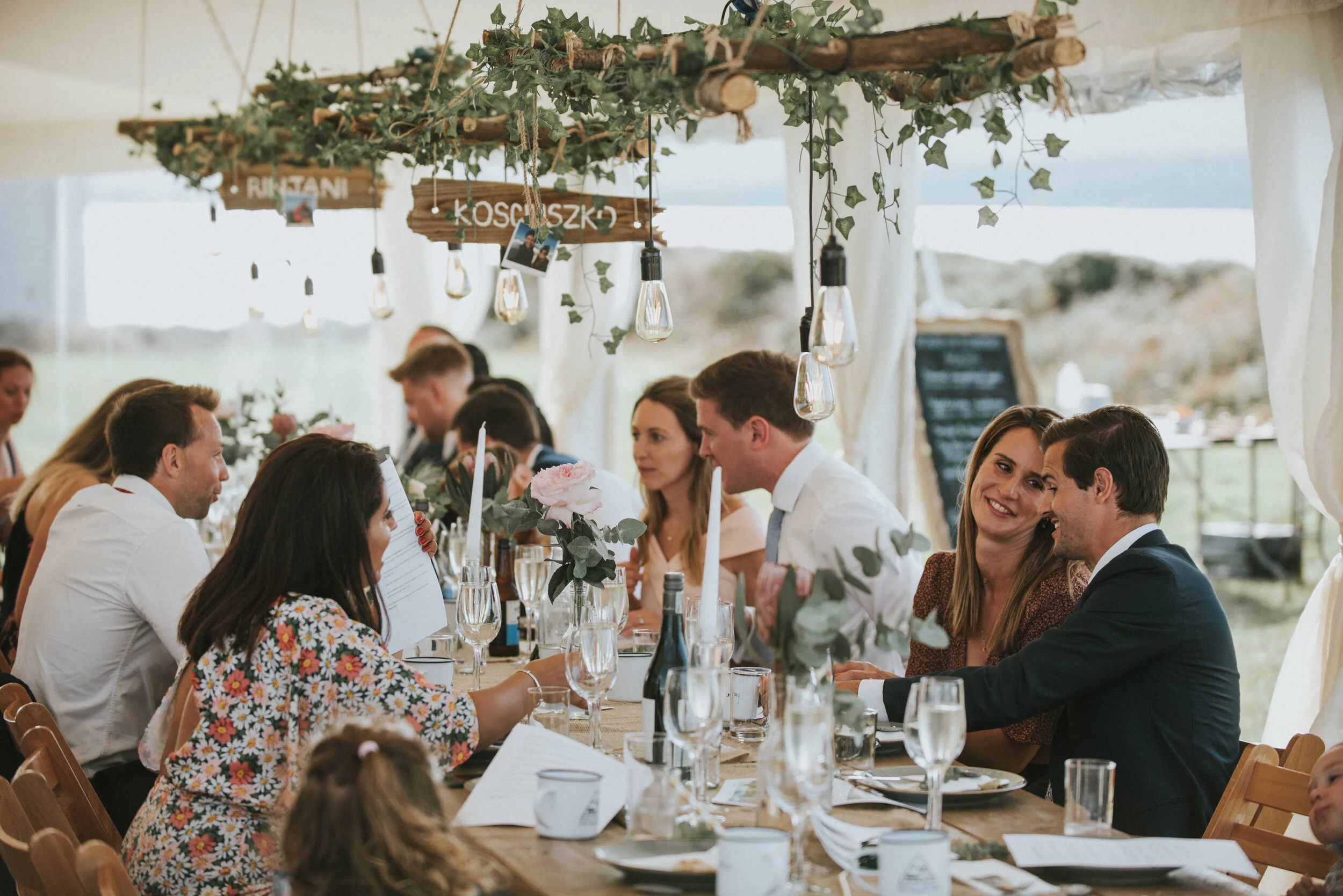 Wedding photo at Greenaway Field in Cornwall. People sitting at a long table during a wedding reception or celebration, decorated with flowers, candles, and hanging lights inside a tent with fabric walls.